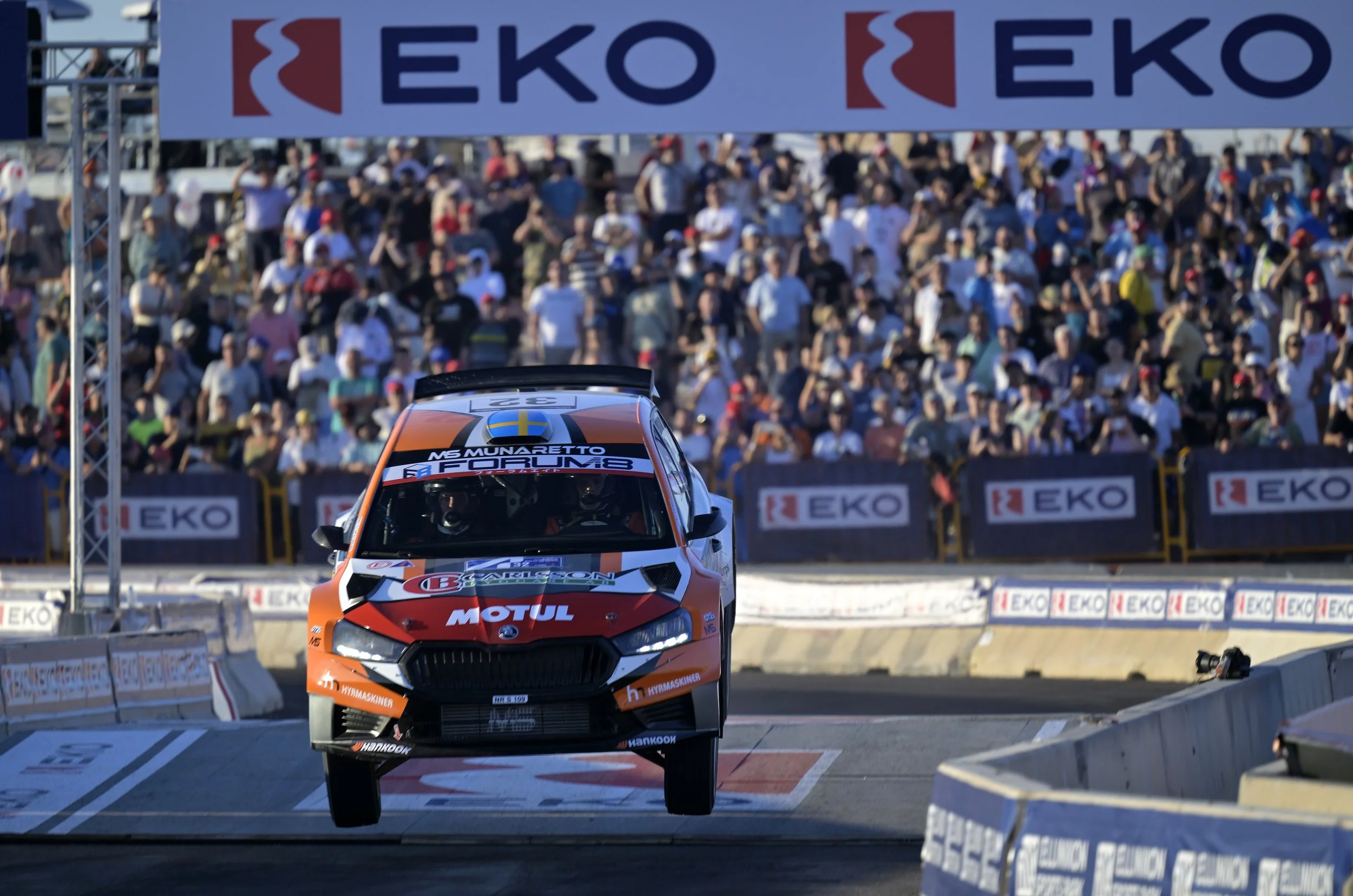 A rally car is jumping off a ramp during a race with a crowd of spectators in the background. The car is orange and black with sponsor logos, and it is mid-air on a dirt track marked by barriers and advertising banners.