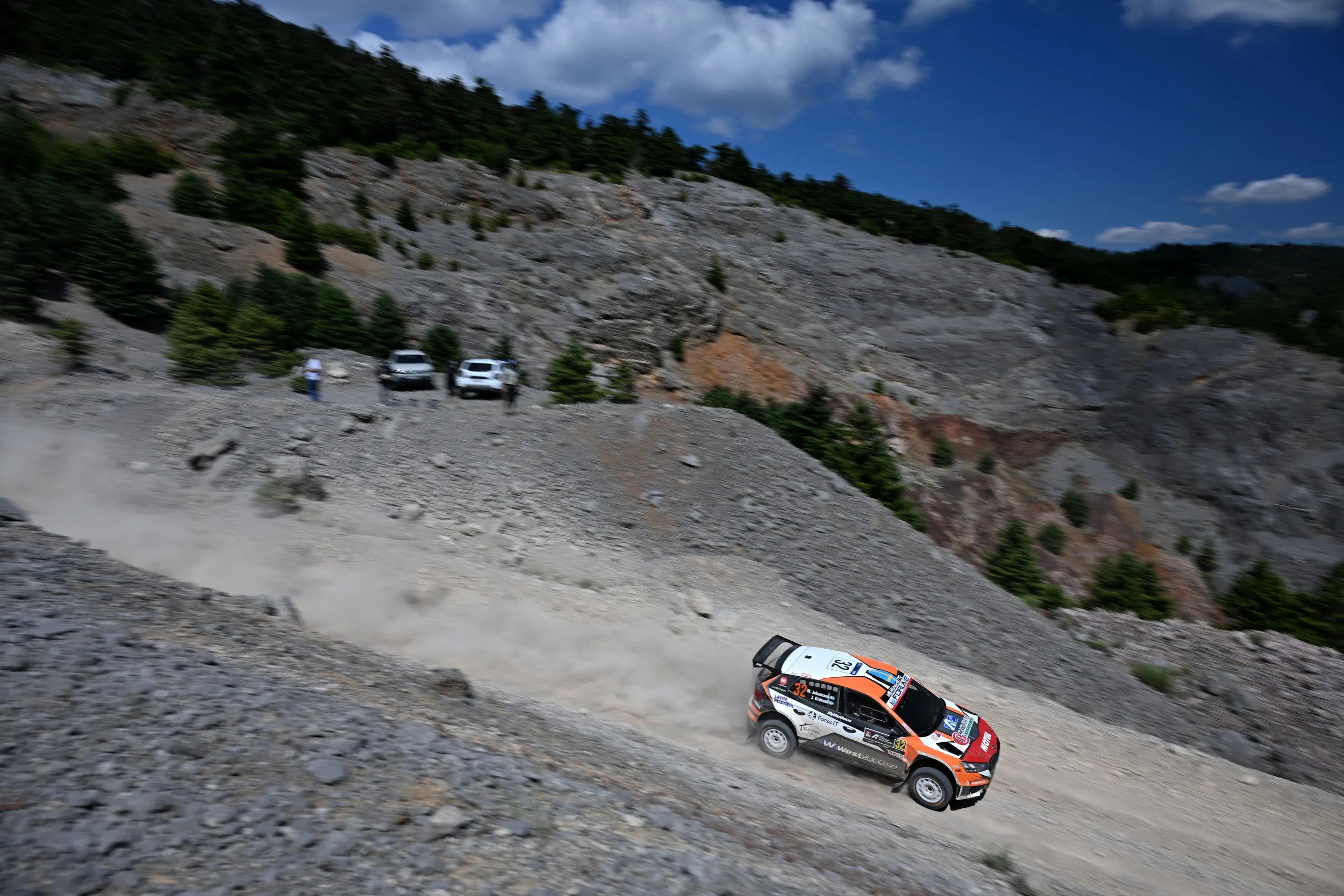 Rally car racing on a dirt mountain road with a steep rocky hillside on the left and a few small trees, under a partly cloudy sky.