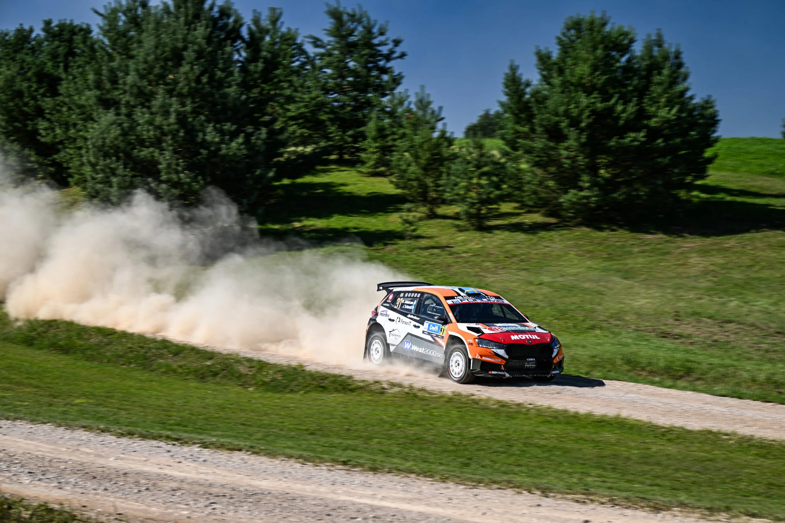 A rally car speeding on a dirt road, kicking up dust, with green trees and blue sky in the background.