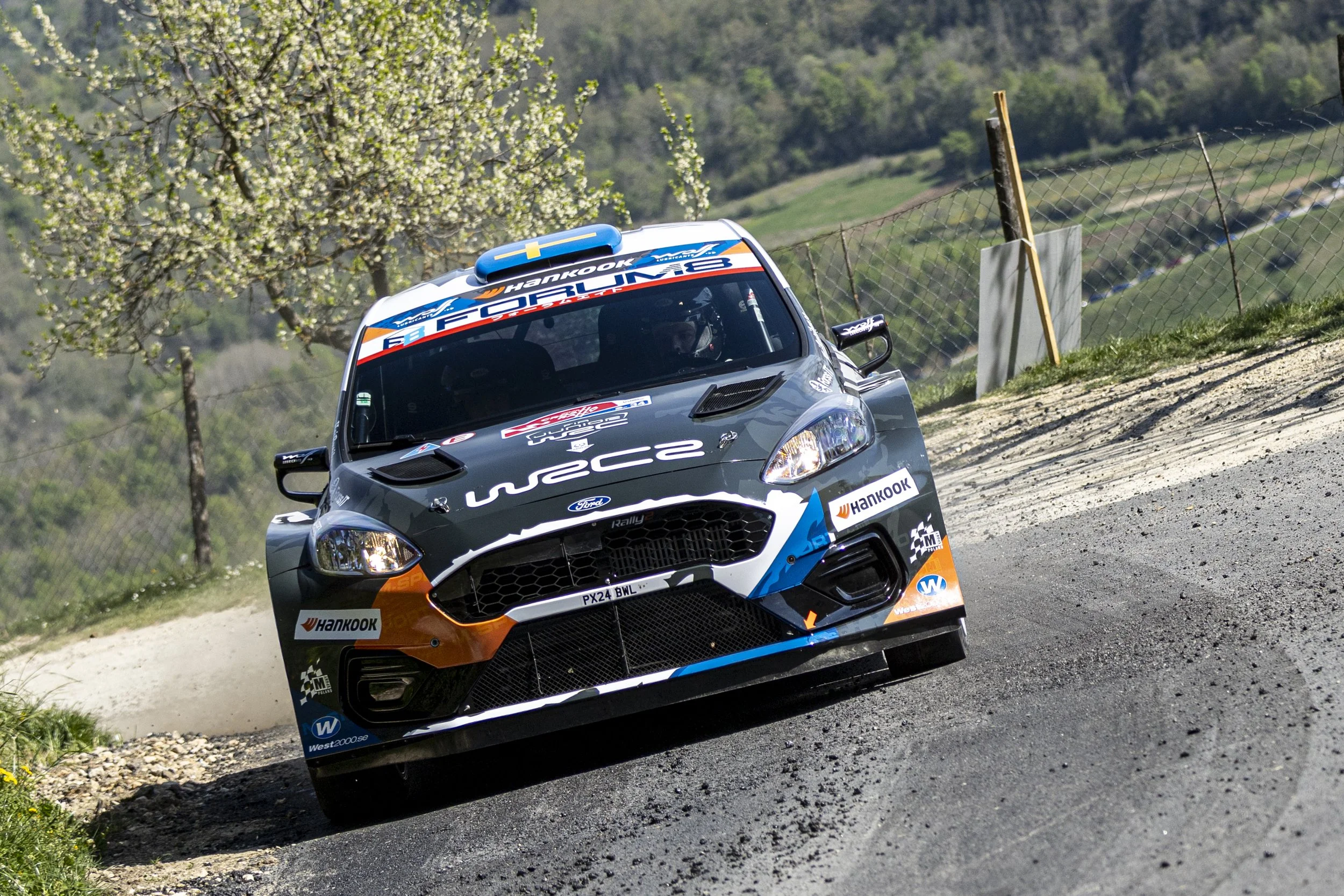 A rally car with Honda and Ford logos racing on a dirt road, kicking up dust, with a backdrop of green hills and trees in spring bloom.