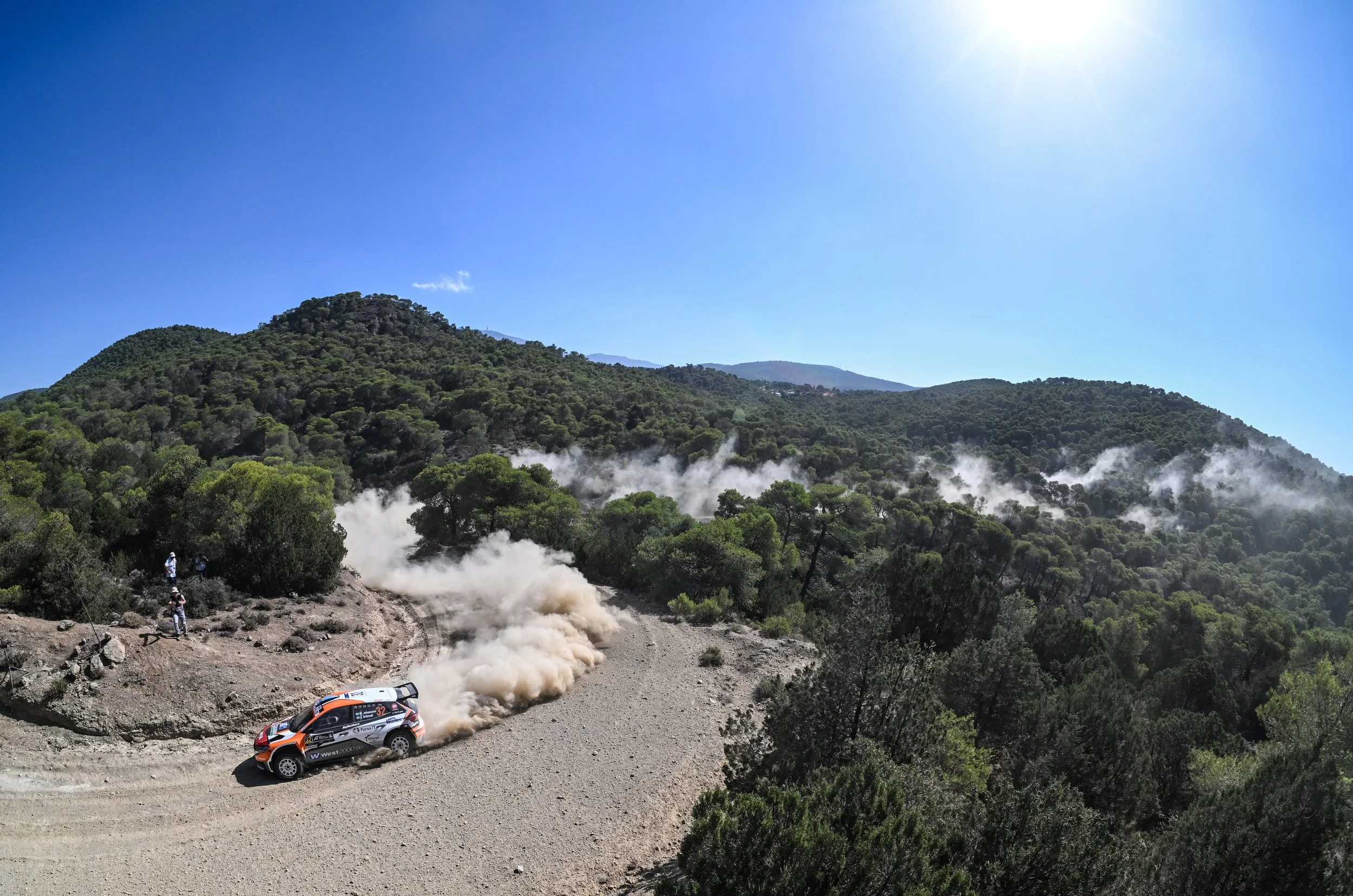 A rally car speeding on a dirt mountain road with dust clouds behind it, surrounded by green trees and hills under a bright blue sky with the sun shining.