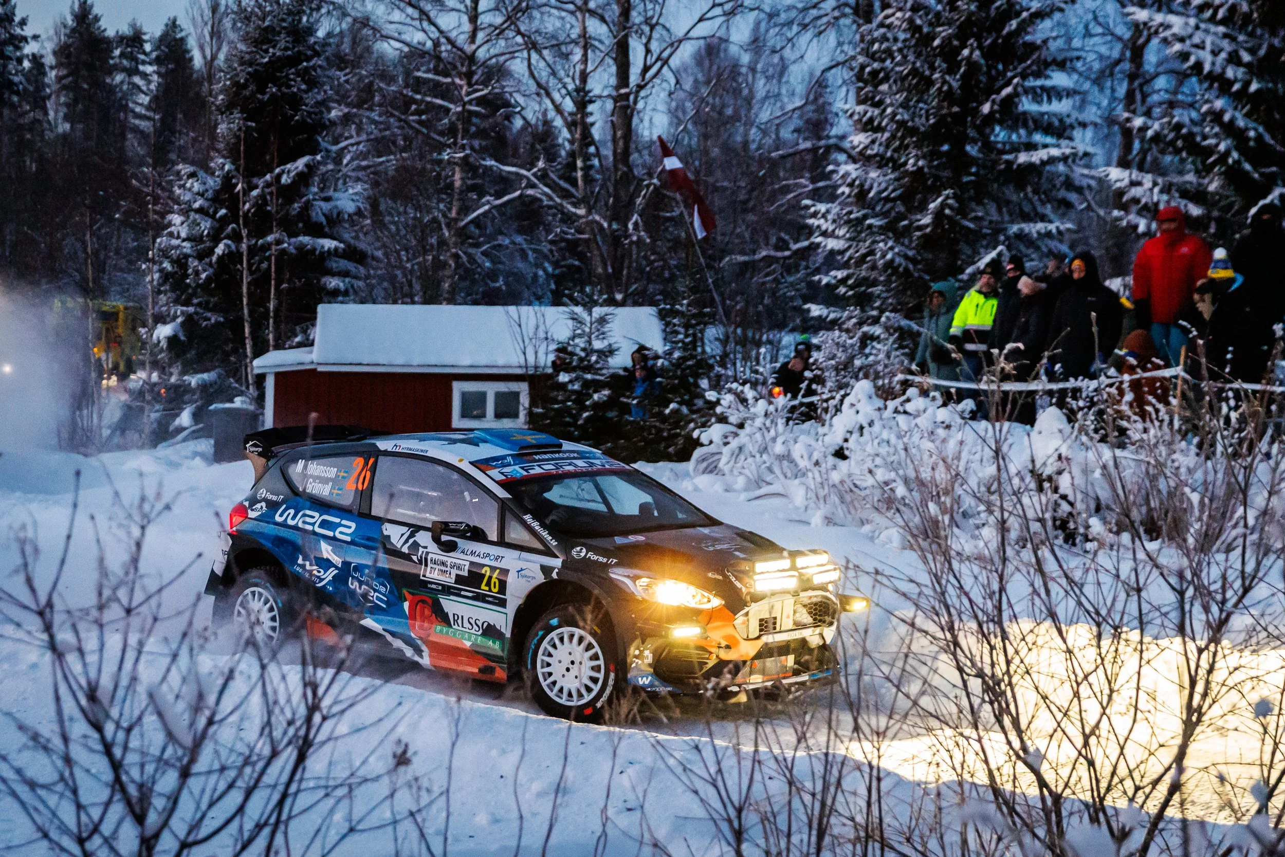 A rally car racing through snow-covered terrain surrounded by trees and spectators during a winter rally event.
