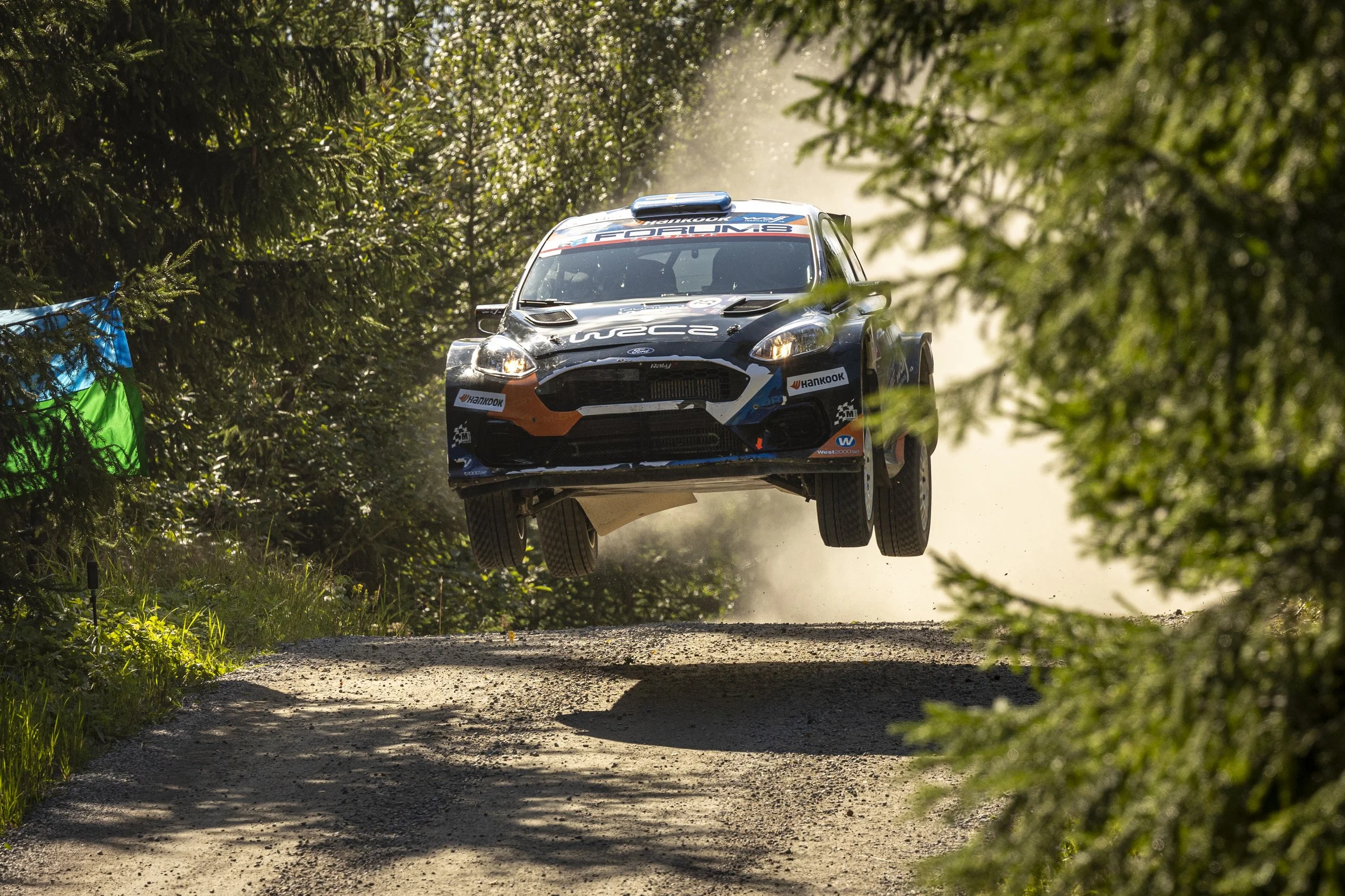 A rally car during a race, airborne over a dirt rally track surrounded by green trees and foliage.