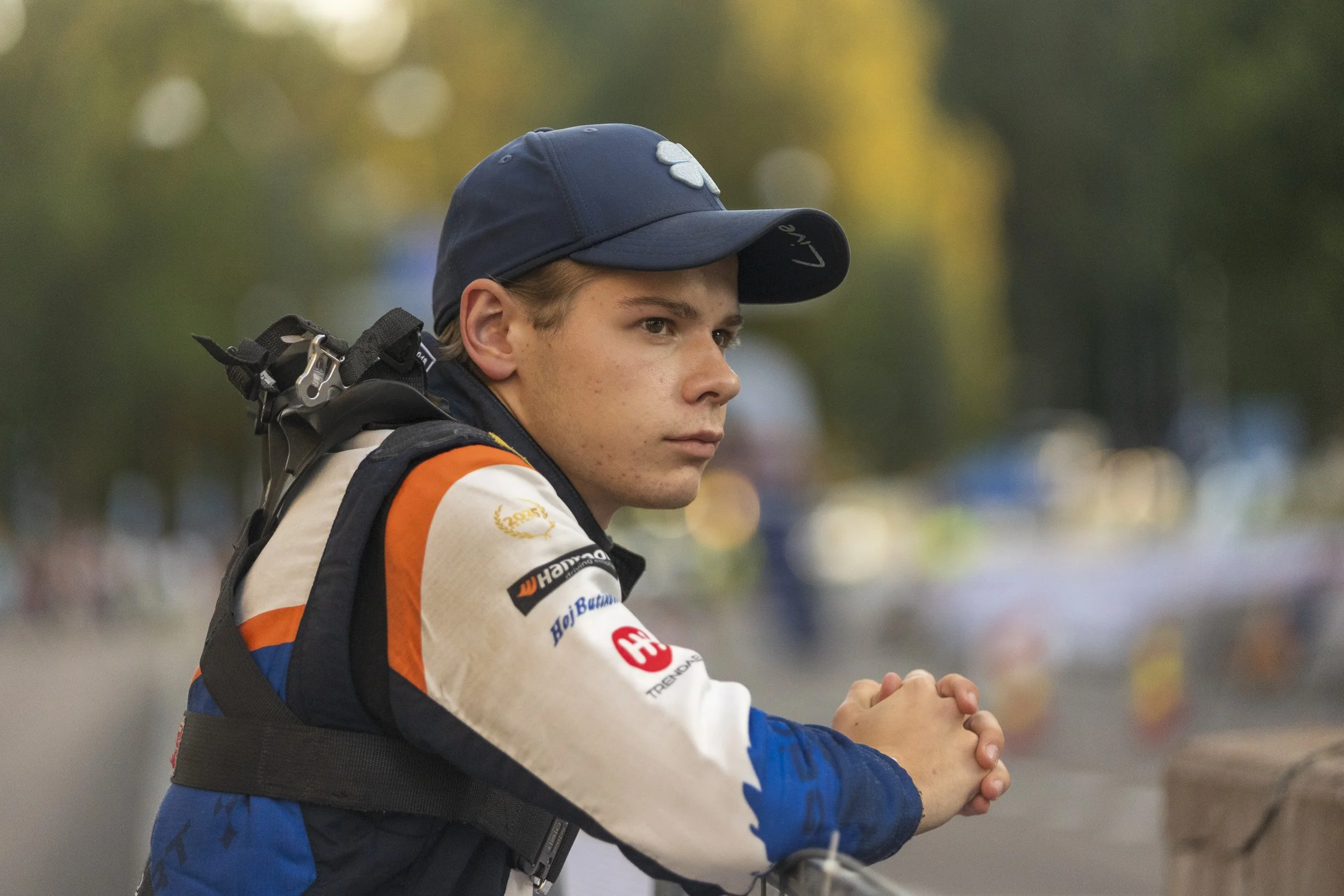 A young male athlete with brown hair, wearing a navy blue cap with a white logo and a racing suit with sponsor logos, holding his hands together in a focused moment.