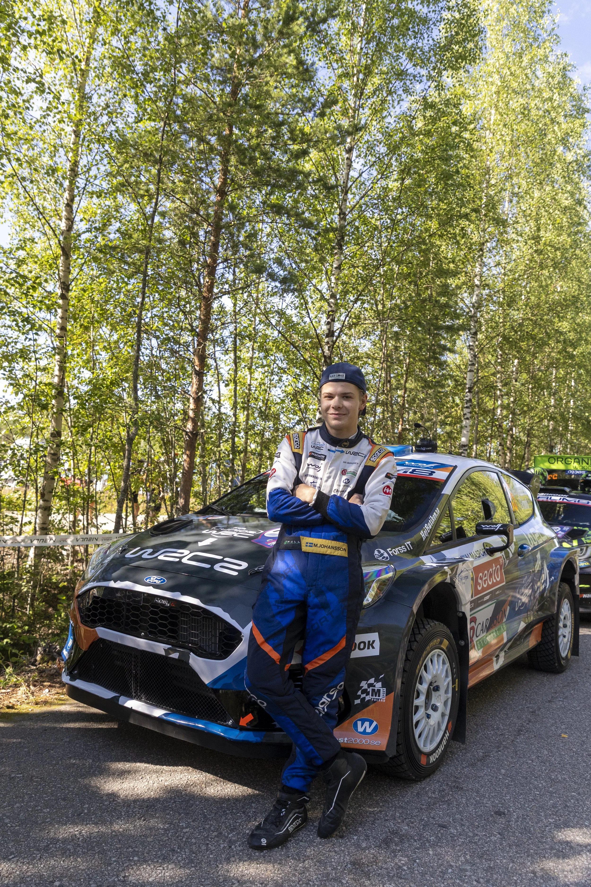 Young male race car driver standing with arms crossed next to a black and blue rally car in a wooded outdoor setting. He is wearing a racing suit, racing shoes, and a baseball cap.