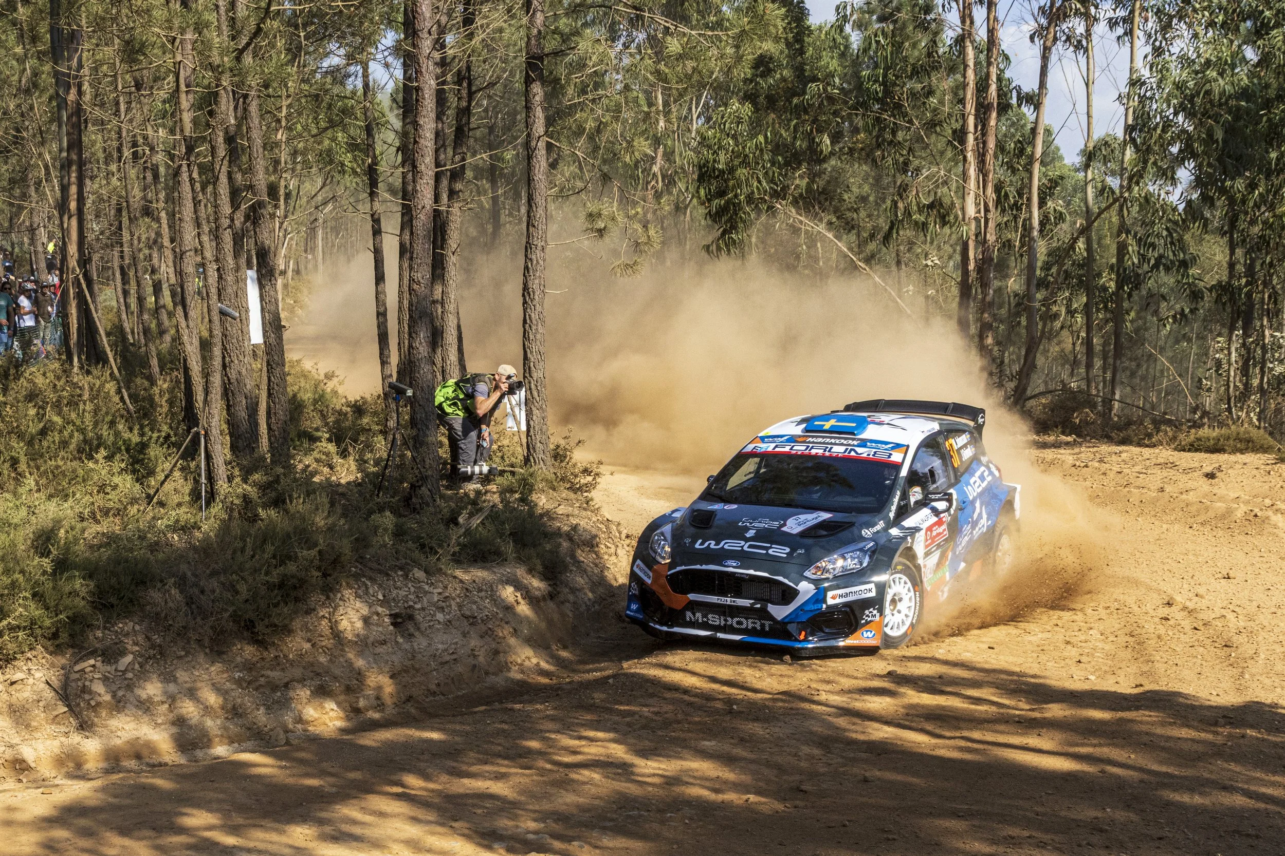 A rally car racing through a dirt forest trail, kicking up dust, with a photographer taking pictures and spectators watching from the side.