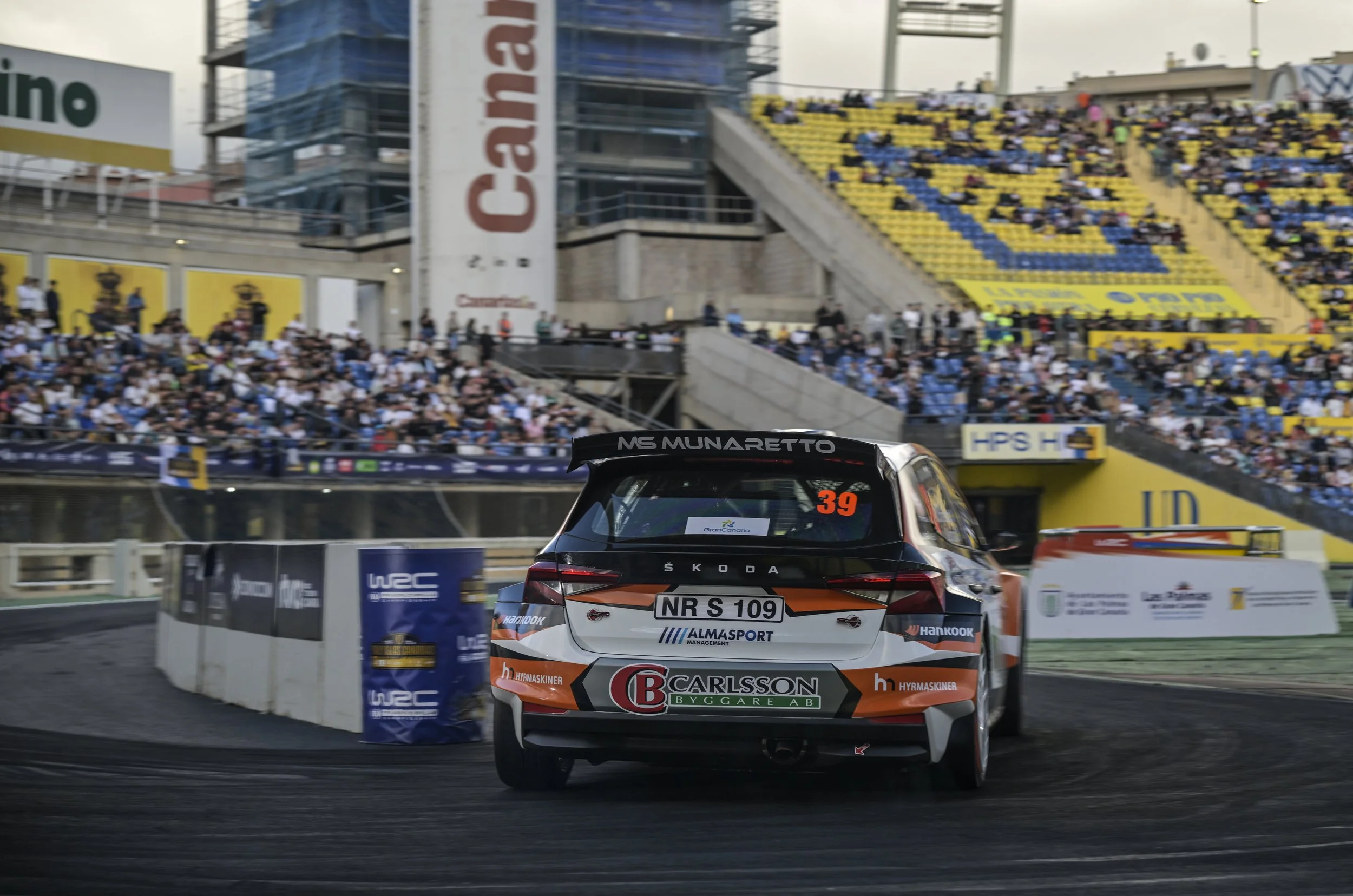 A race car on a track at a racing event with spectators in the stands and advertisements in the background.