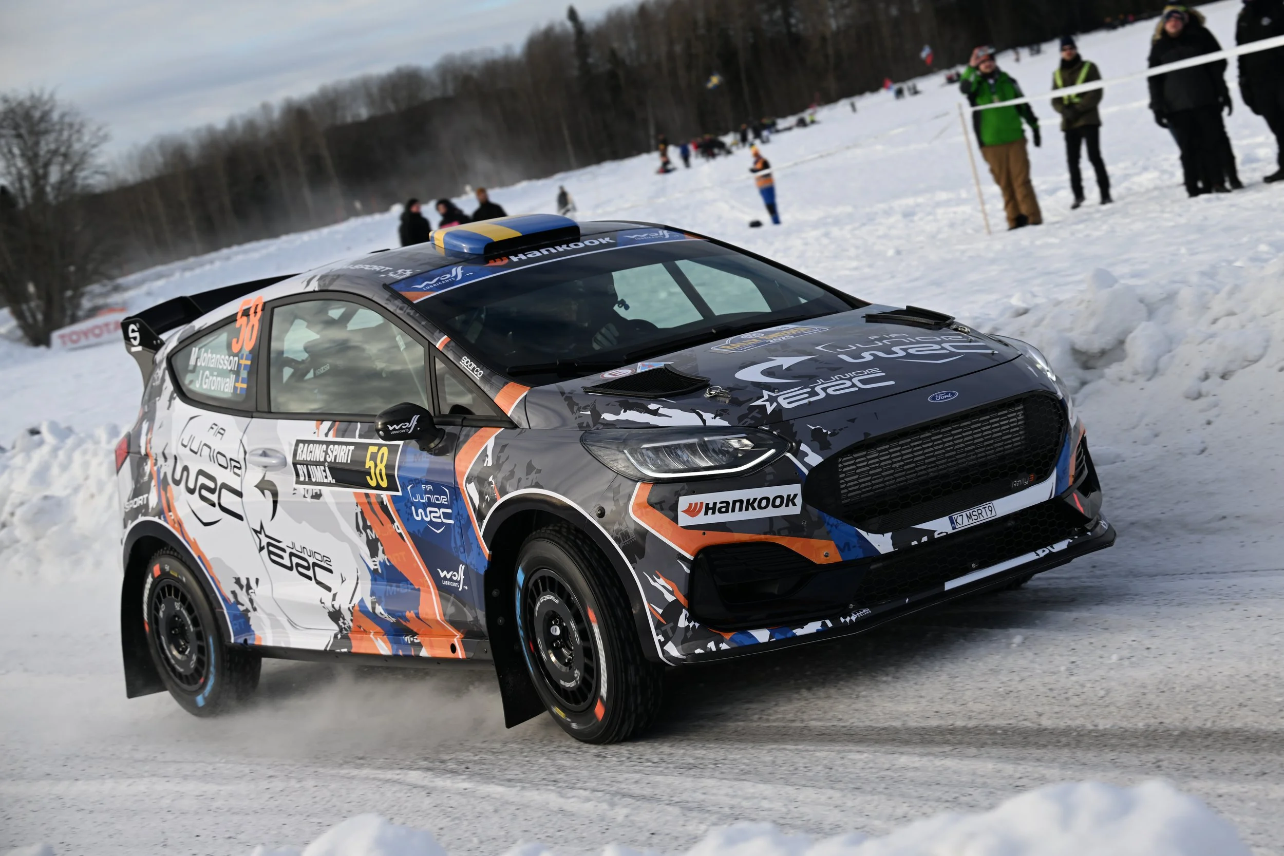 A race car driving on a snowy winter road during a racing event, with spectators watching behind a safety barrier in the background.