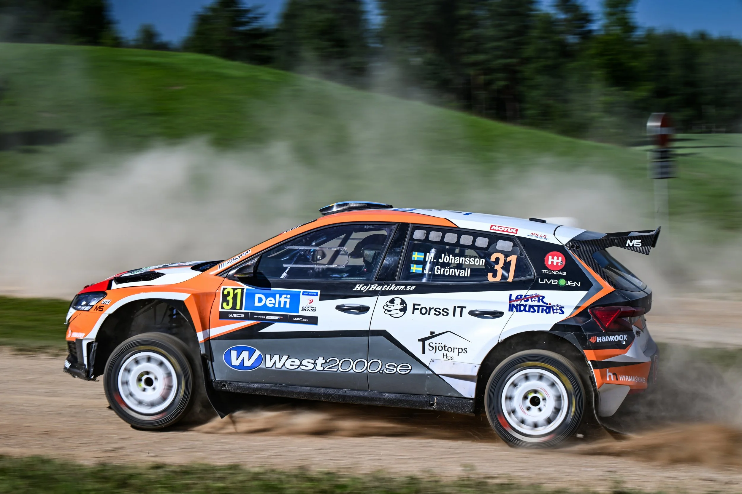 A rally car driving on a dirt road with dust behind it, surrounded by trees and grass.
