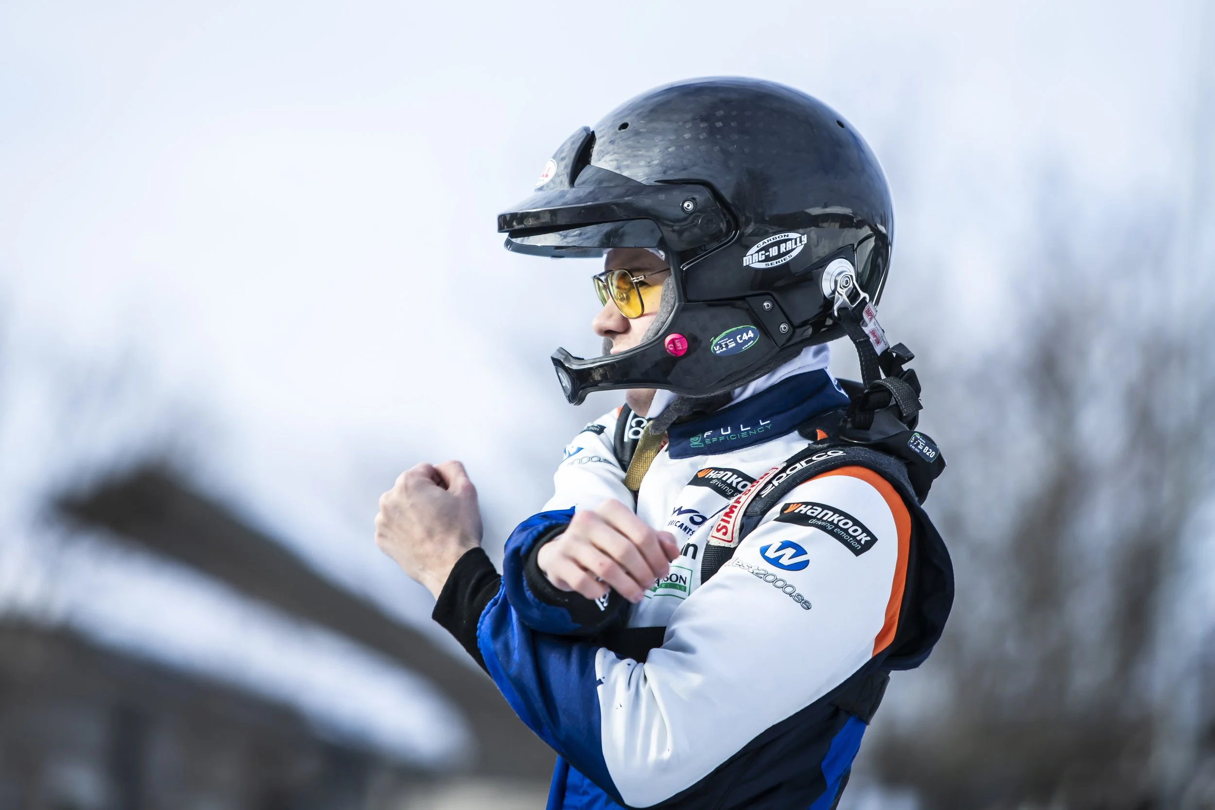 Person in racing gear wearing a black helmet and sunglasses, crossing arms, standing outdoors with snowy background.