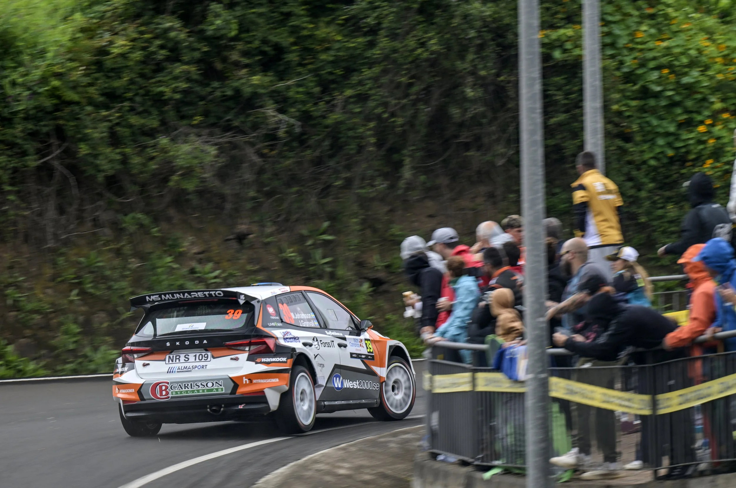 A rally car racing on a curved road with a crowd of spectators standing behind a barrier, against a backdrop of green trees.