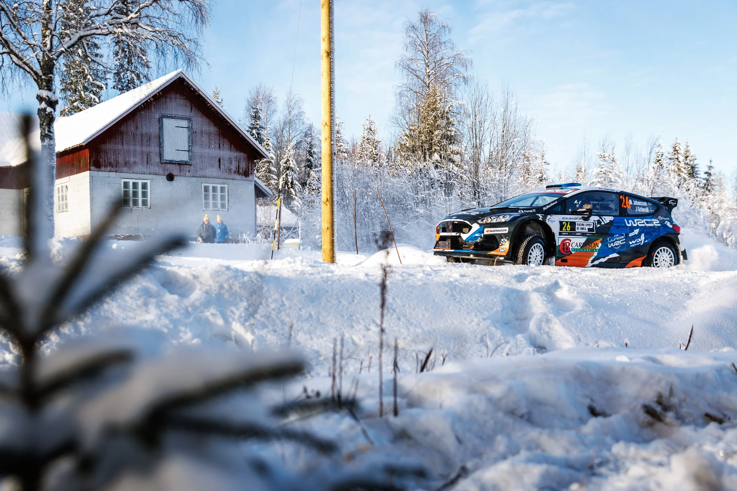 A snowy landscape with a house on the left, trees covered in snow, a yellow utility pole, and a black rally car with racing decals on the snow-covered ground.
