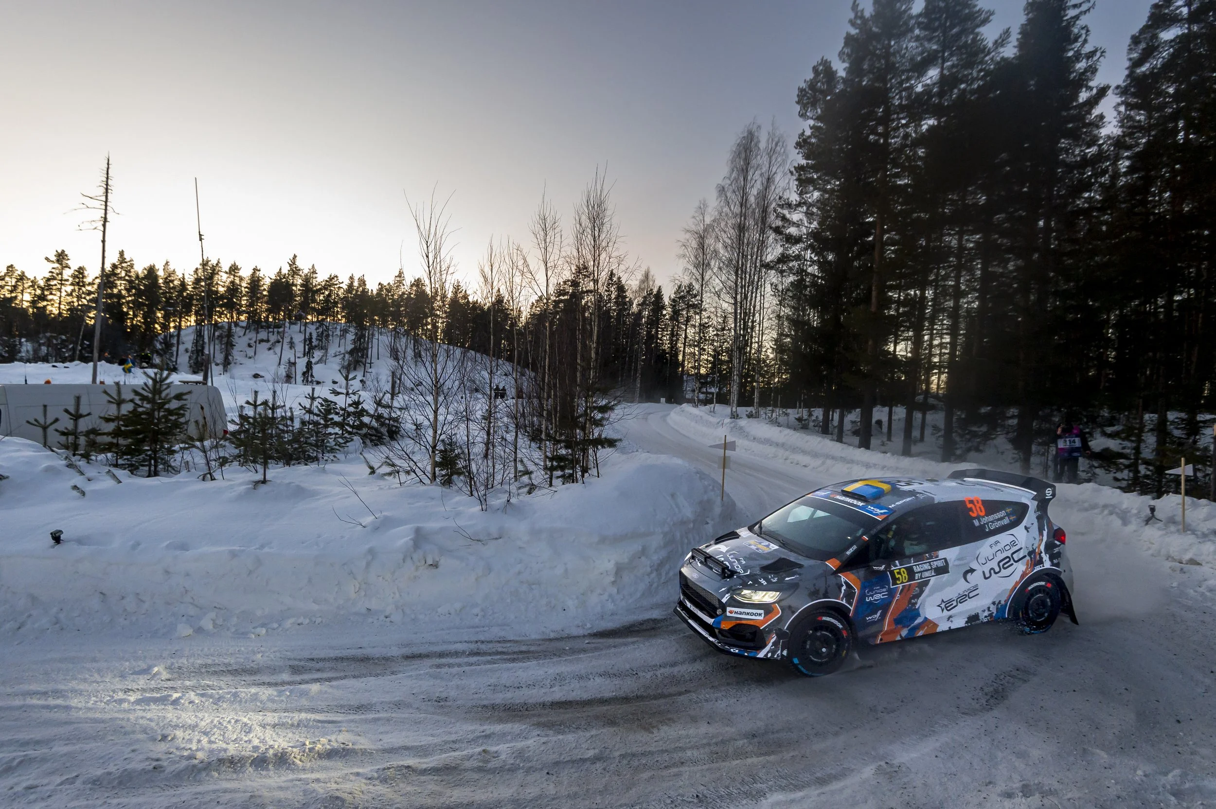 A rally car racing on a snowy road in a winter forest setting, with a backdrop of tall trees and a clear sky during late afternoon or early evening.
