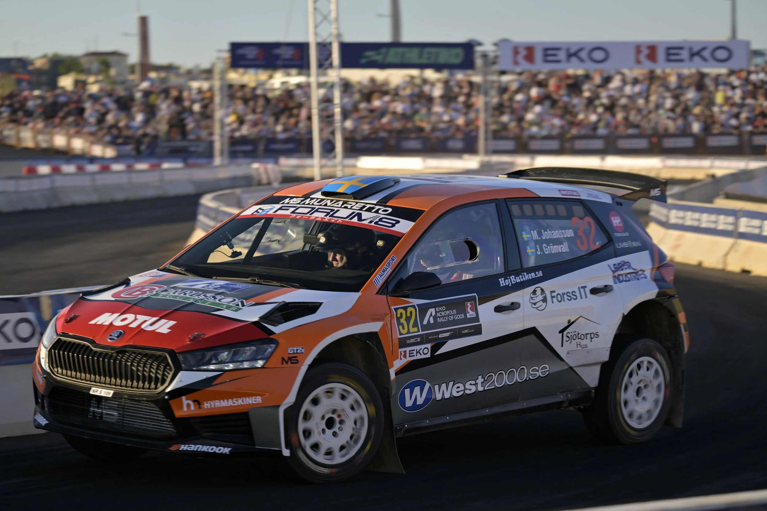 A rally car racing on a track with a crowd watching from the stands in the background.