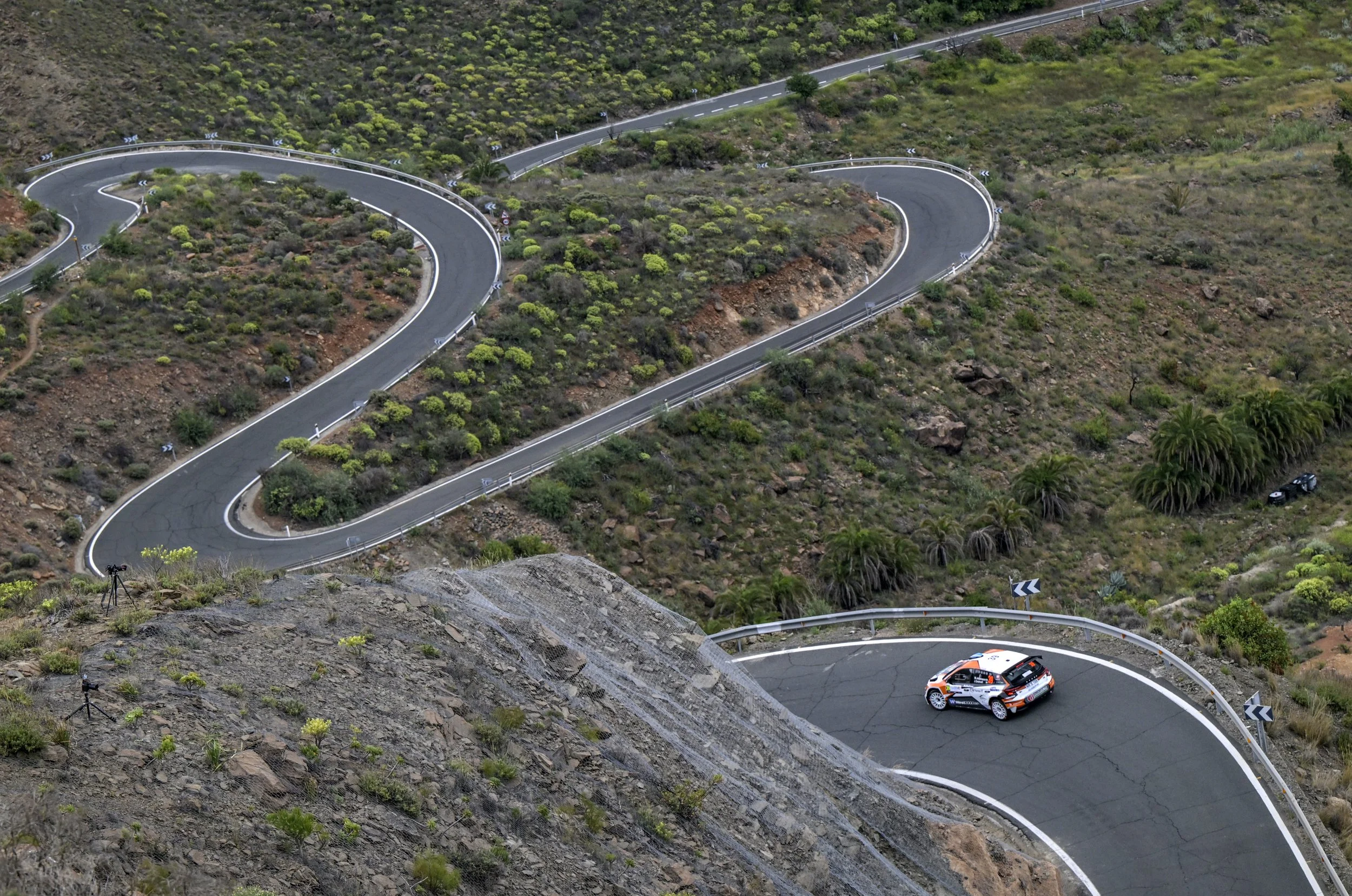 A winding mountain road with sharp turns, guardrails, and a rally car racing through the curves surrounded by green vegetation and rocky terrain.