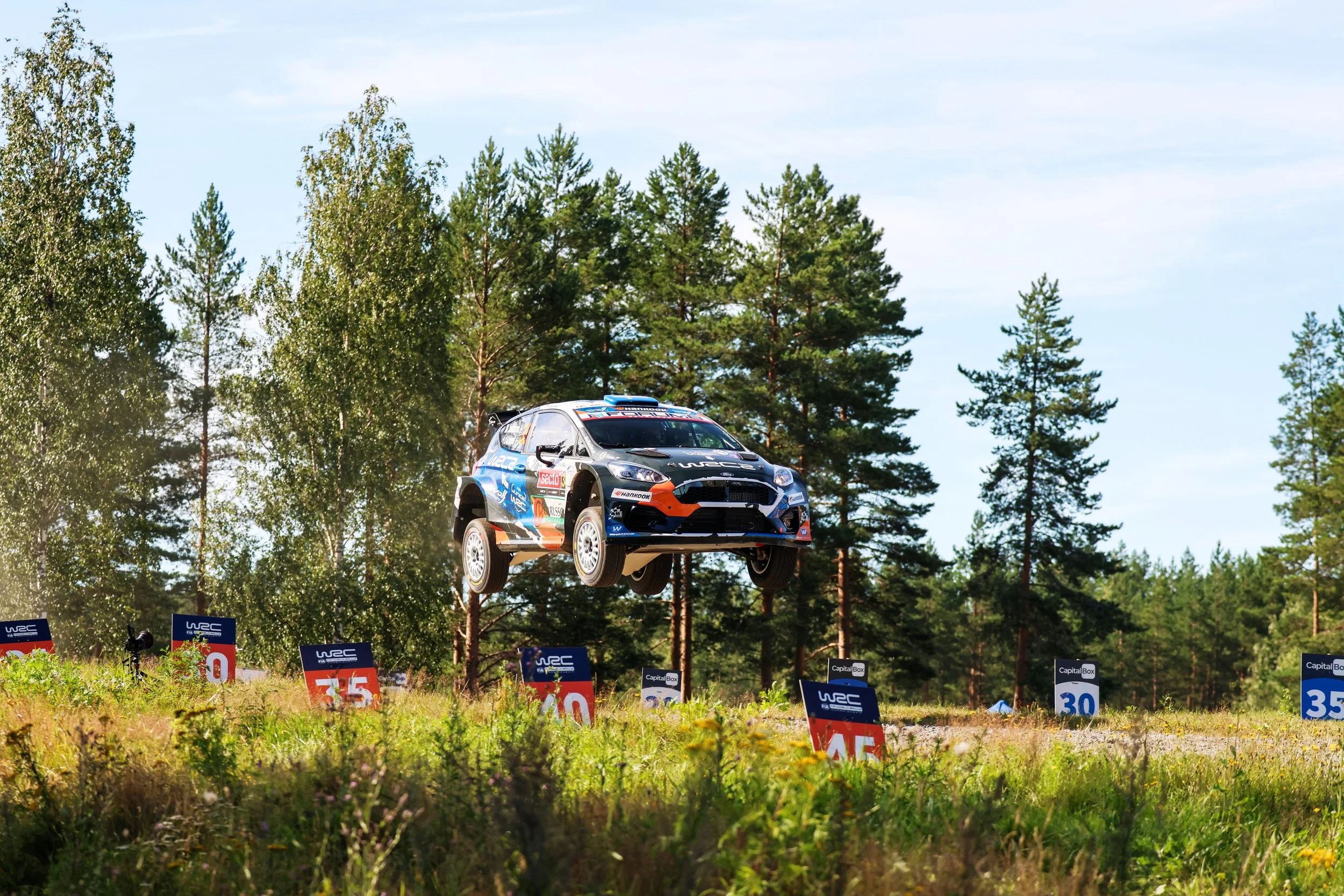 A rally car is mid-air jumping over a bump on a dirt track surrounded by trees and rally signs.