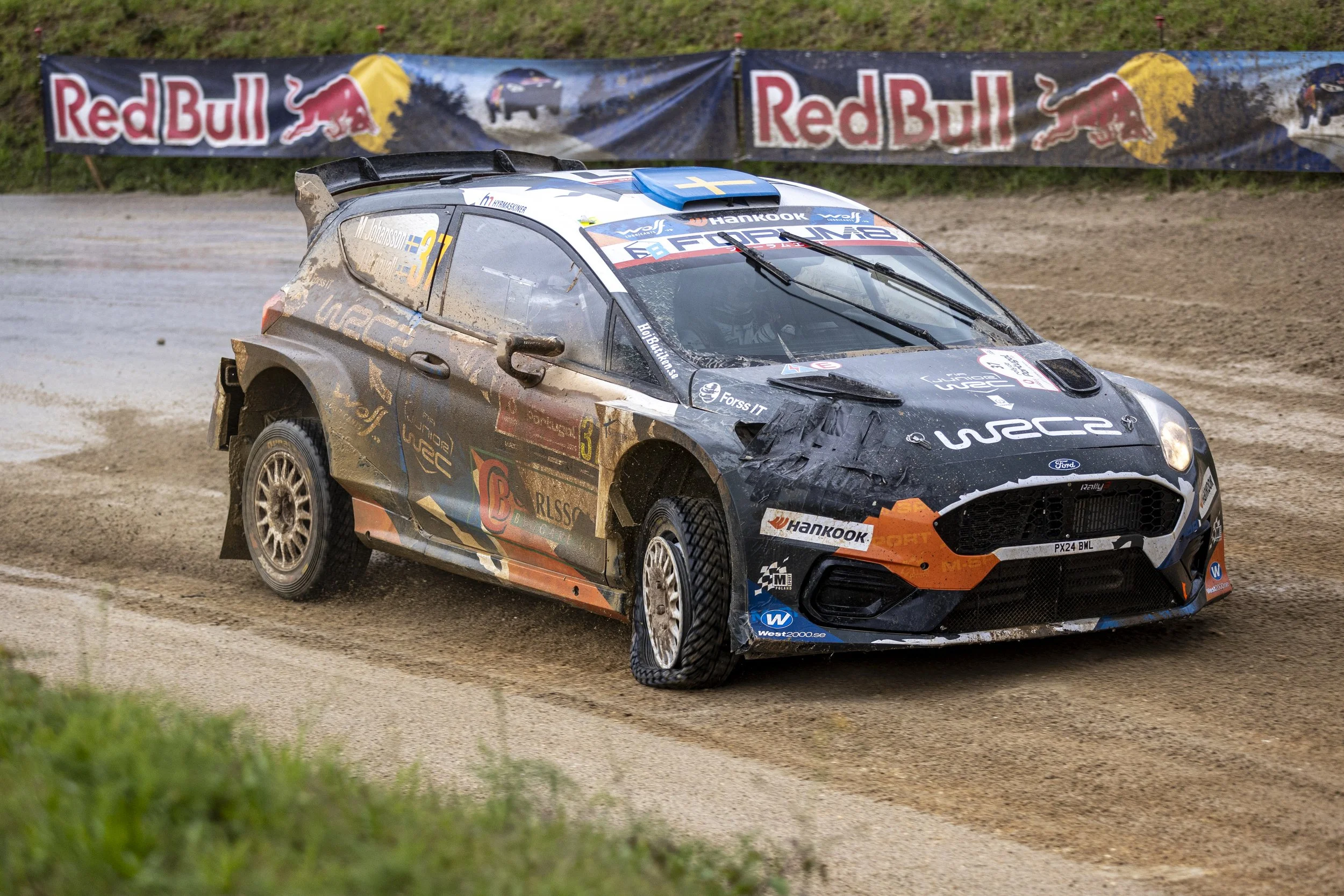 A rally car driving on a dirt track with mud splattered on its sides, during a race with Red Bull banners in the background.