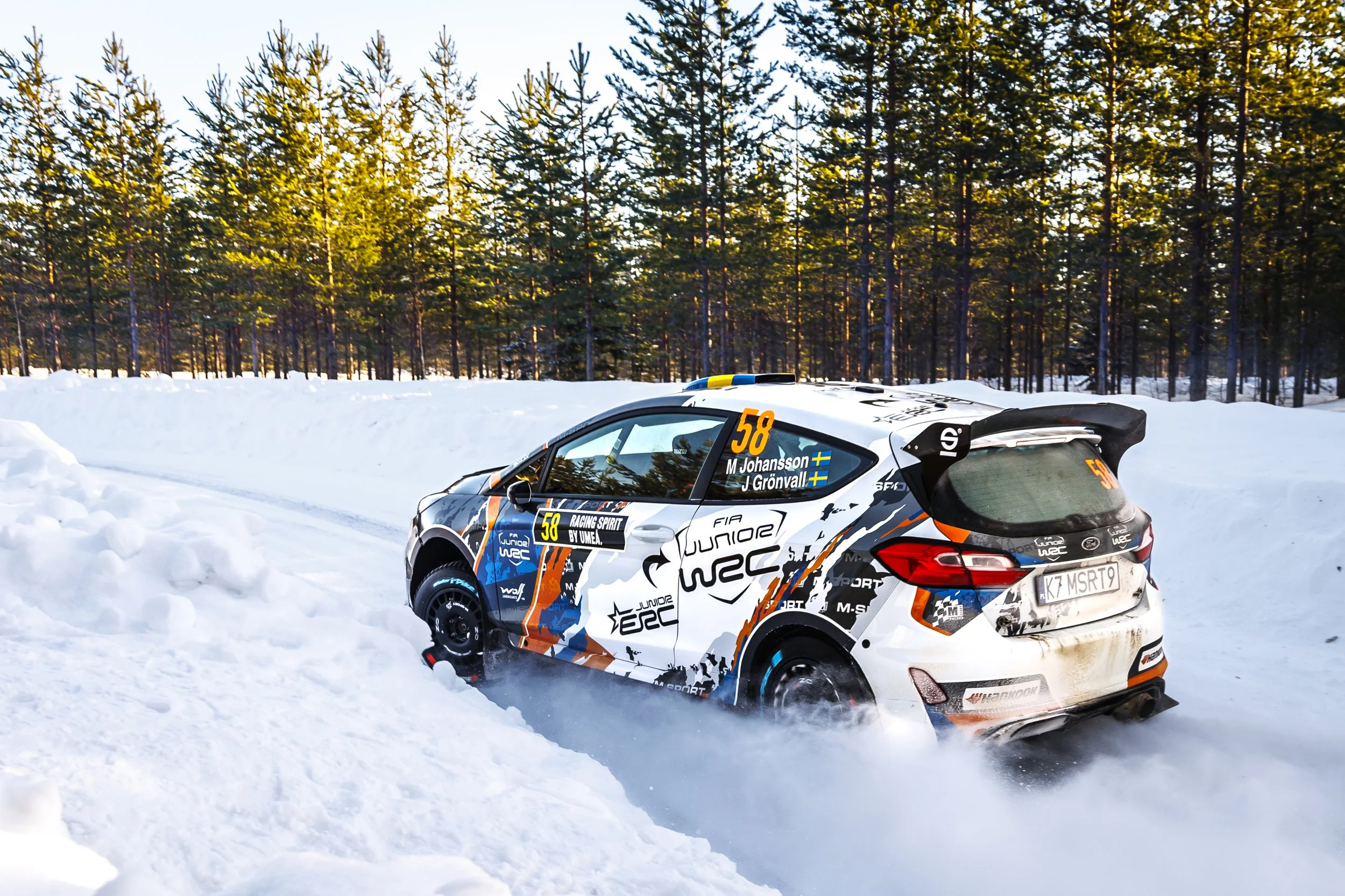 A rally car speeds through snow, creating a spray of snow behind it, with a background of tall pine trees and a clear sky.