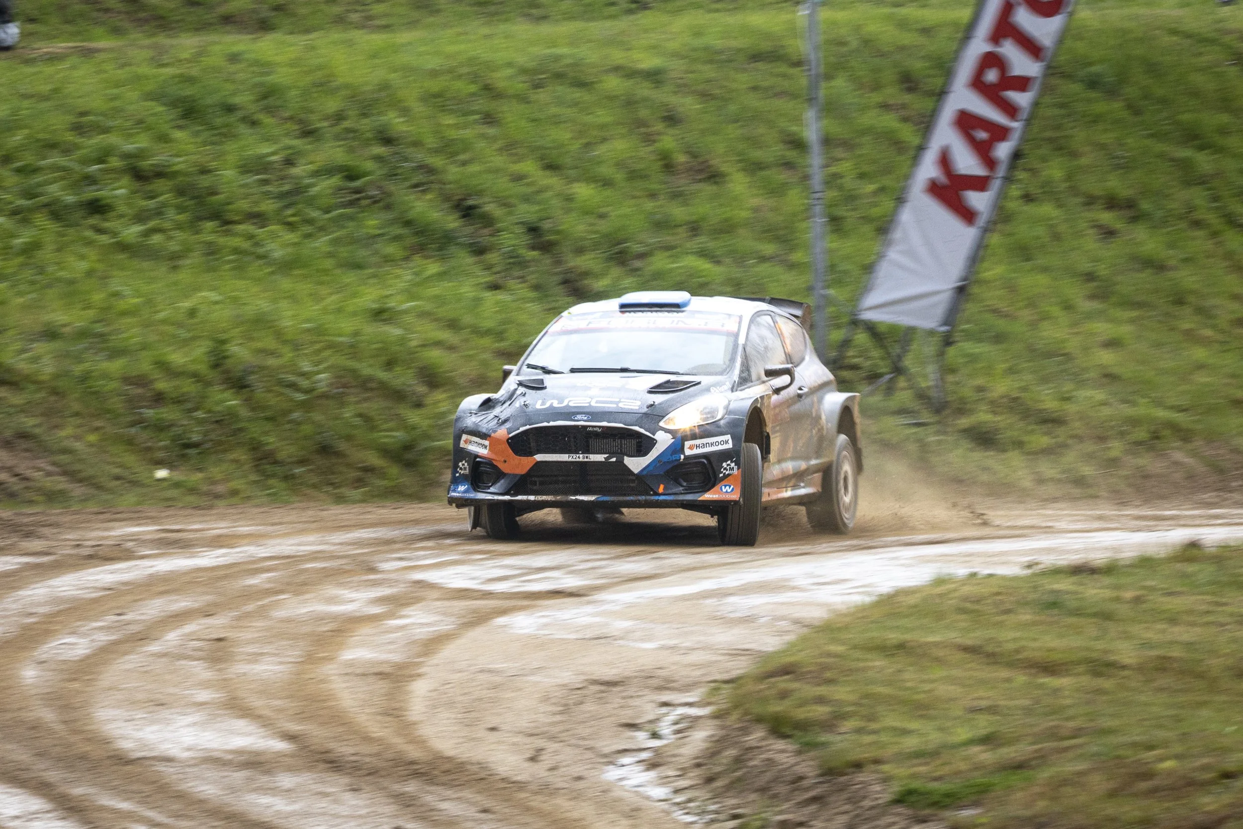 A rally car racing on a dirt track with green grass and hill in the background, kicking up dust behind it, and a large red and white banner along the side of the track.