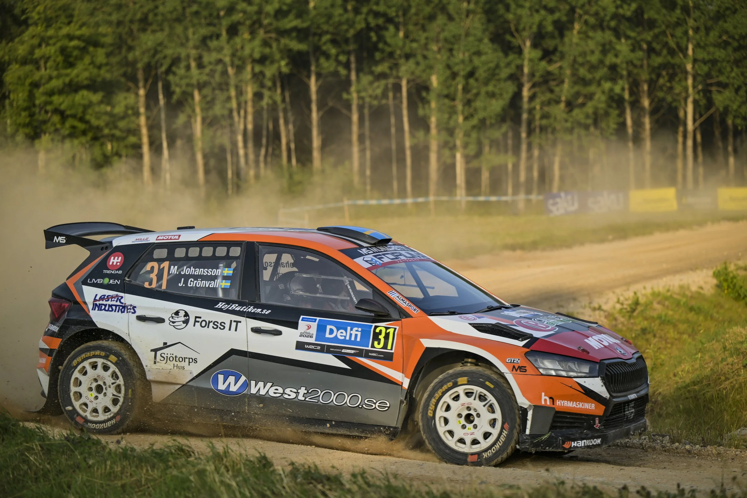 Rally car racing on dirt road with dust and trees in the background.