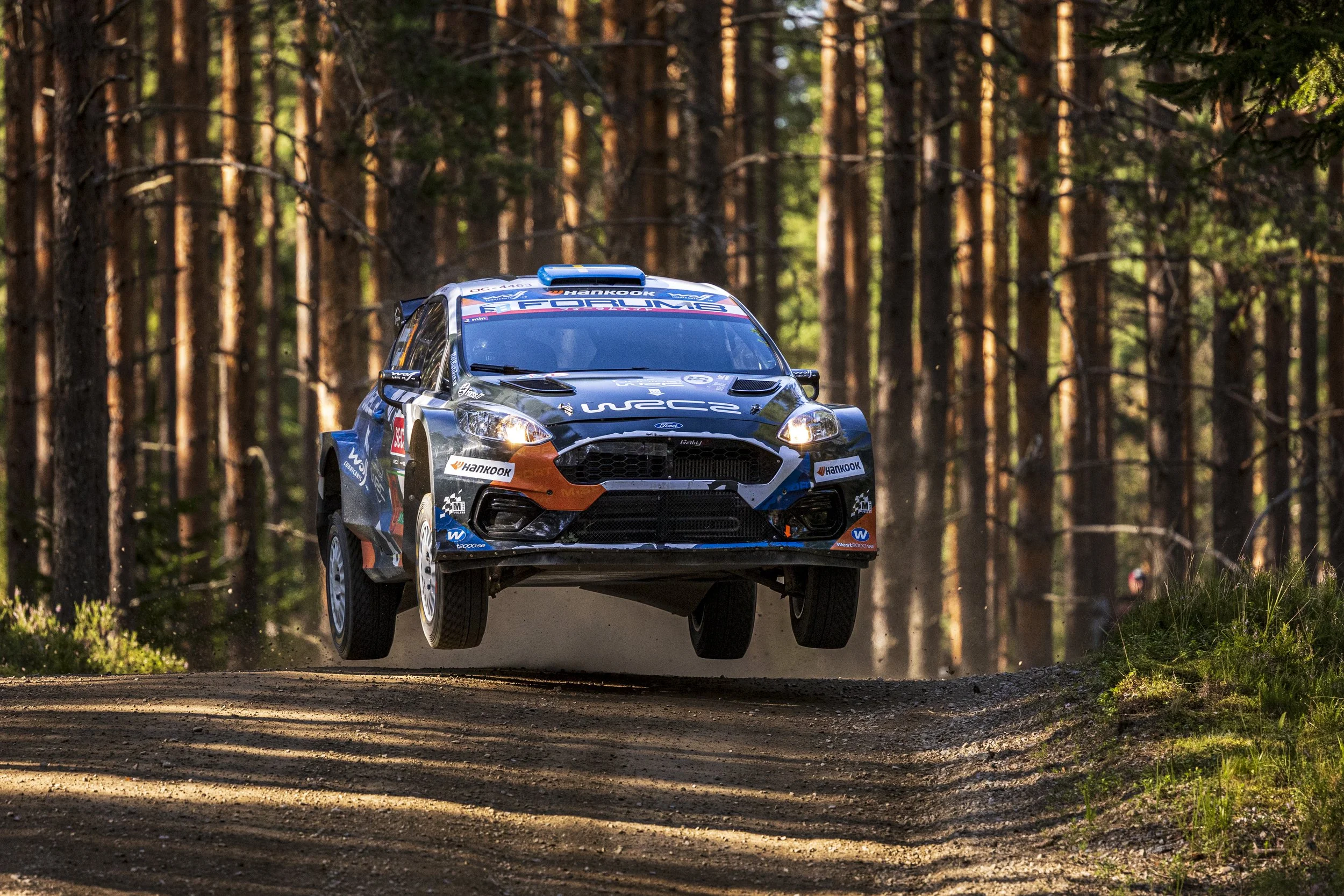 A rally car flying through the air while racing on a dirt road in a dense forest of tall pine trees.