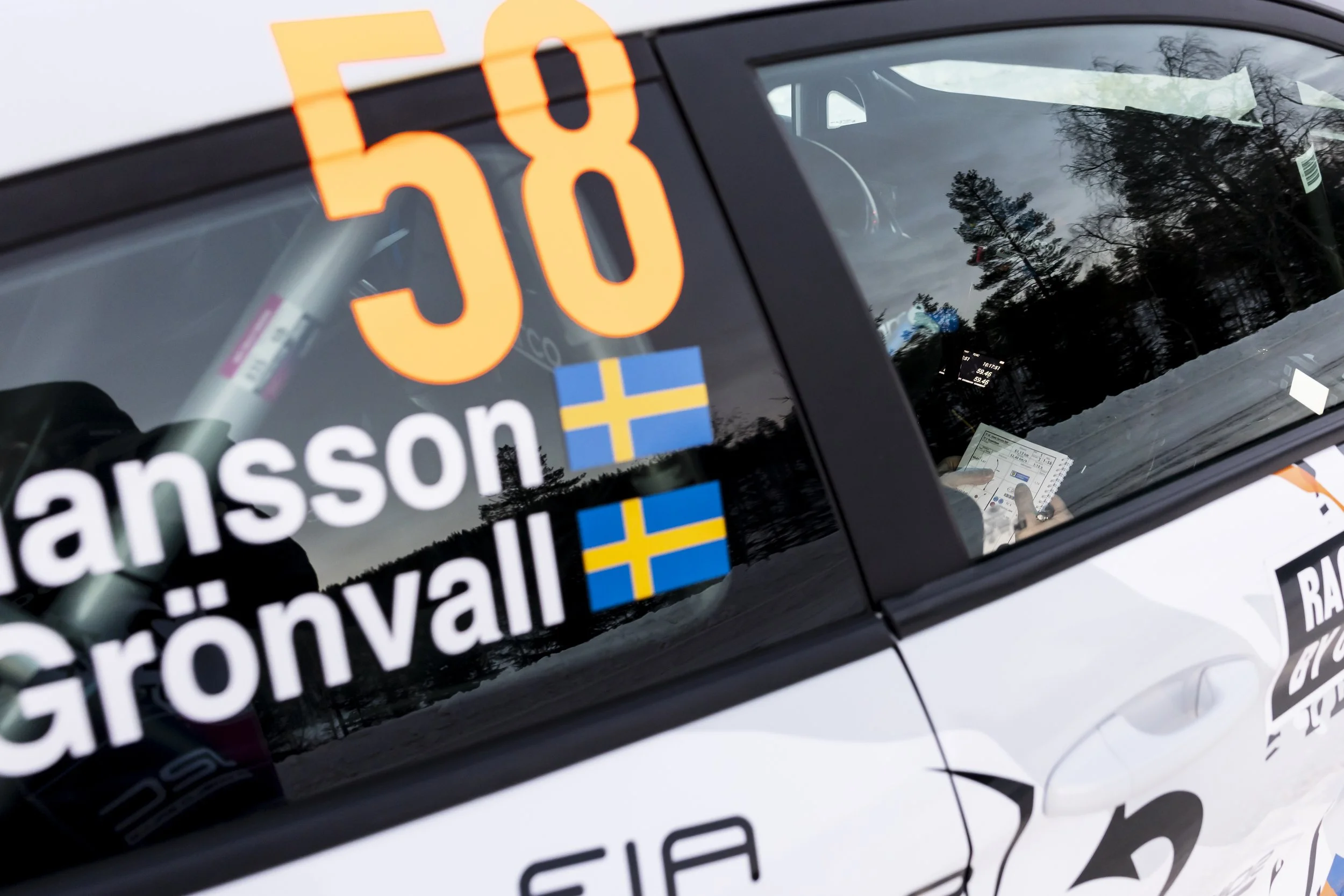 Close-up of a vehicle window with stickers, including a large orange number 50, Swedish flags, and partially visible text in Swedish, reflecting trees and the cloudy sky outside.
