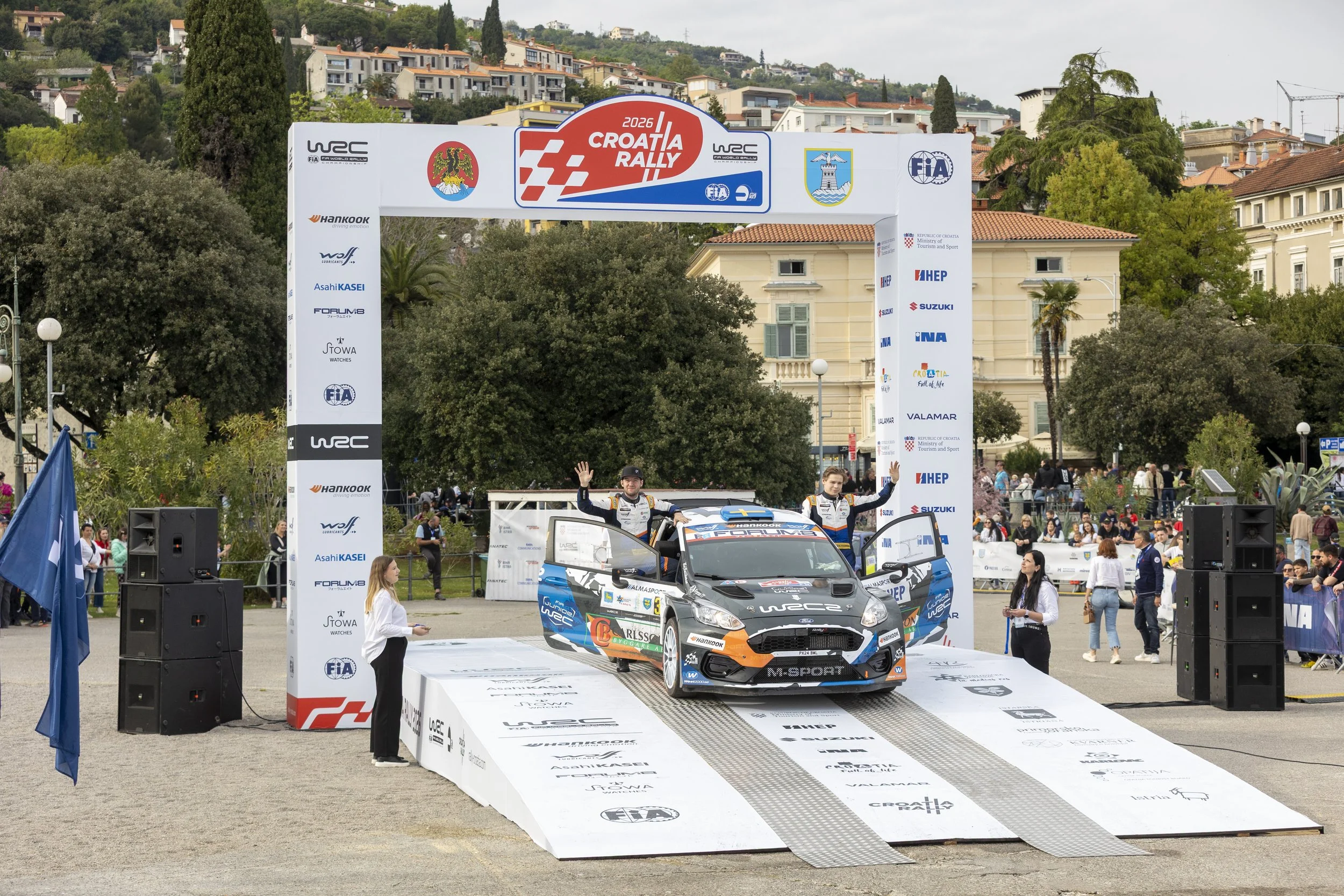 Race car on a winners' podium at the 2026 Croatia Rally with rally drivers celebrating and waving, surrounded by a crowd and banners with sponsor logos.