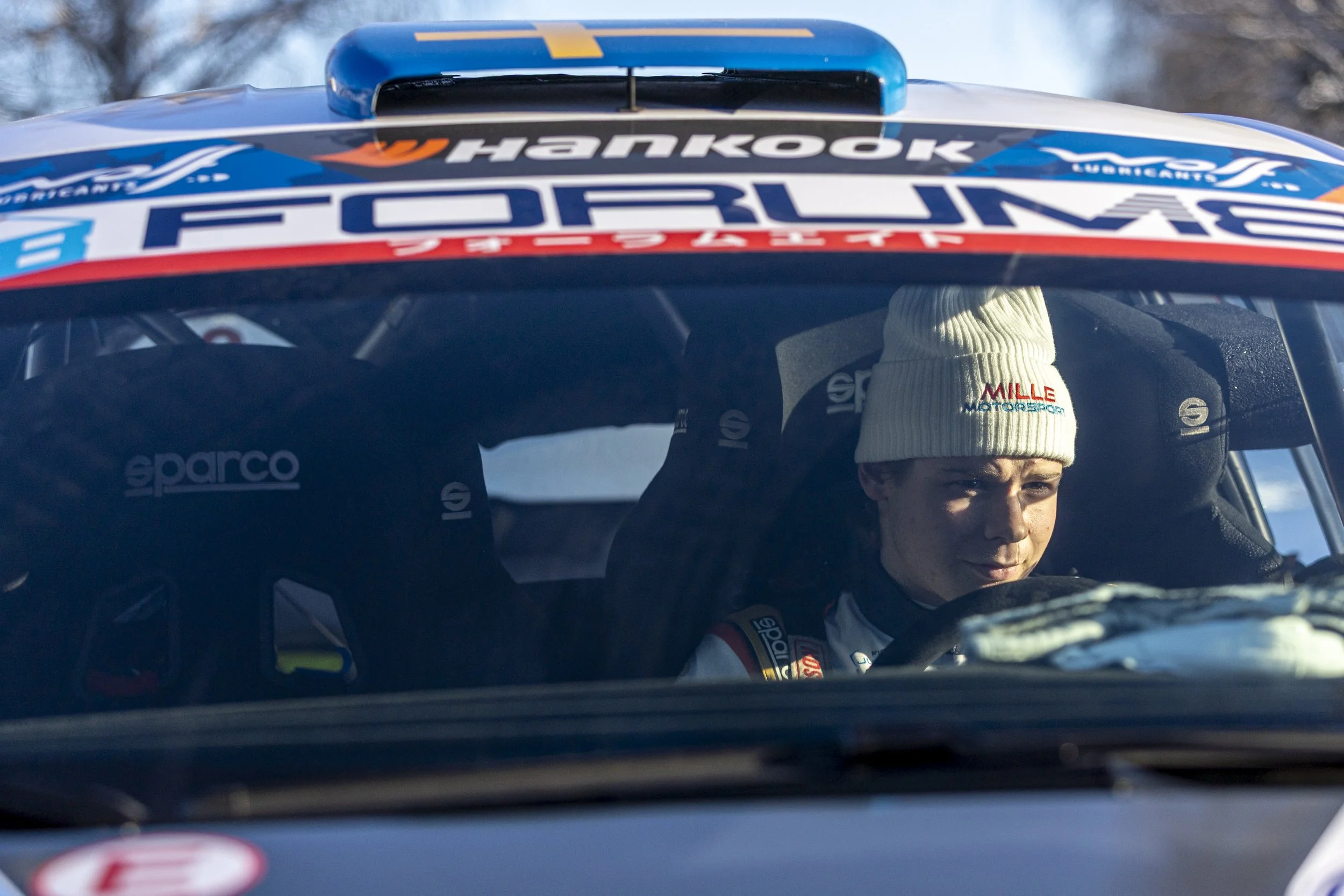 A race car driver wearing a white beanie and racing suit sitting in a colorful rally car with sponsor logos, including Hankook and Forlam, behind the windshield. The background shows a winter outdoor setting.