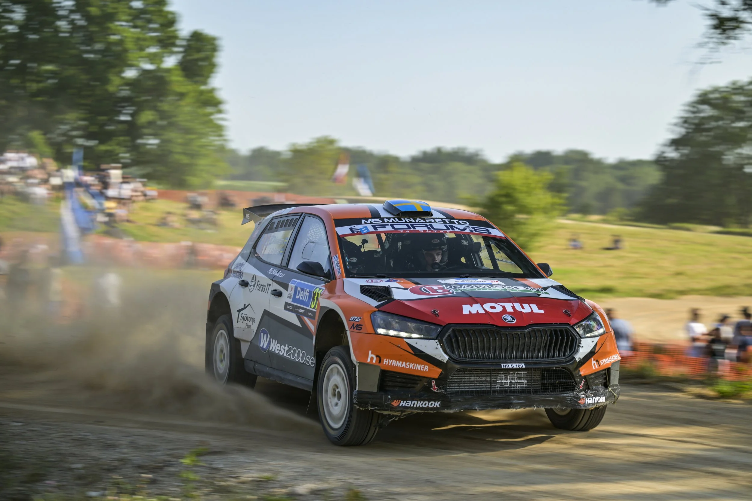 A rally car racing on a dirt track with trees and spectators in the background.