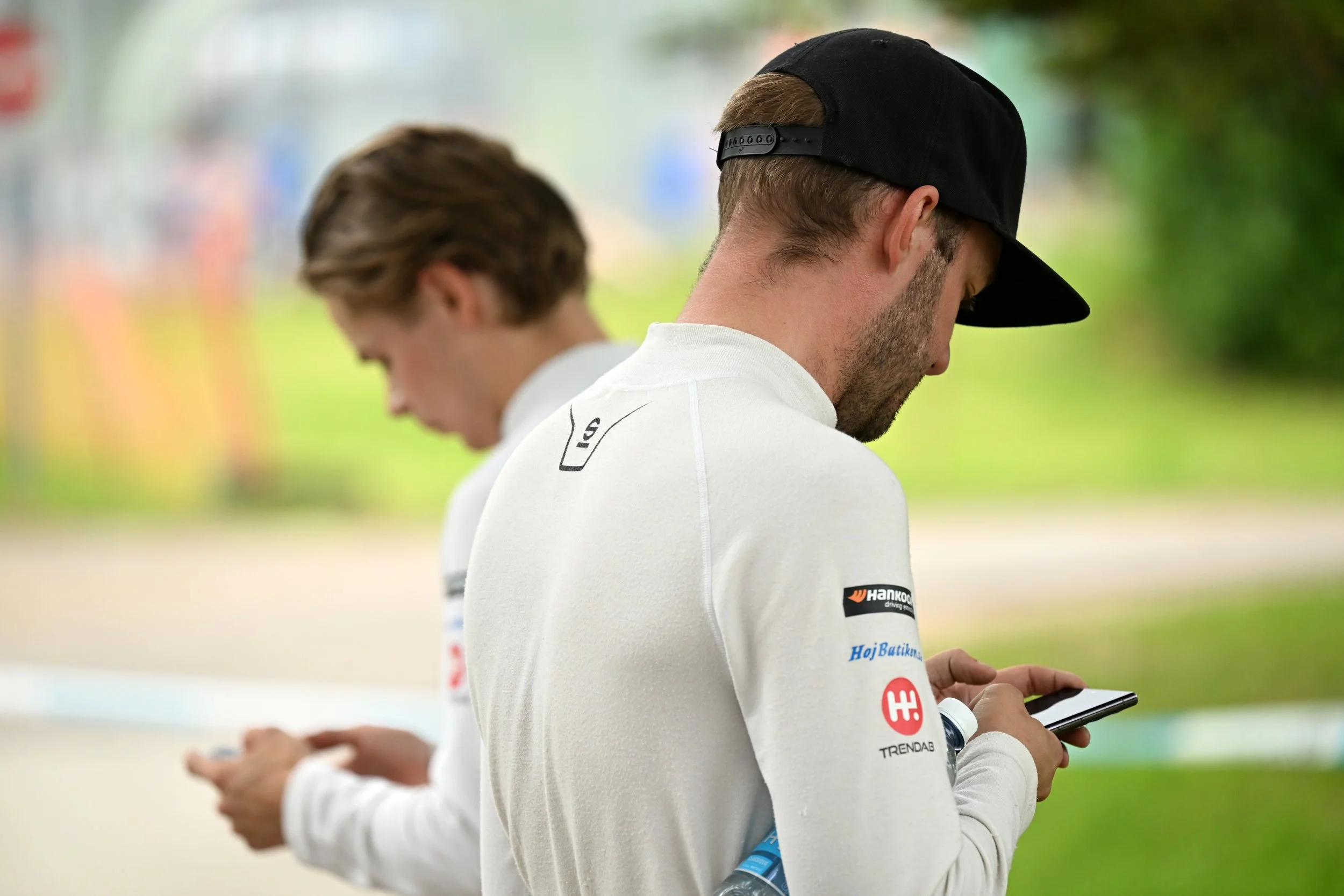 Two young men standing outdoors, looking at their phones. The man in the foreground has a black cap worn backwards and a white long-sleeve shirt with sponsor logos. The background is blurred with green trees and colorful signs.
