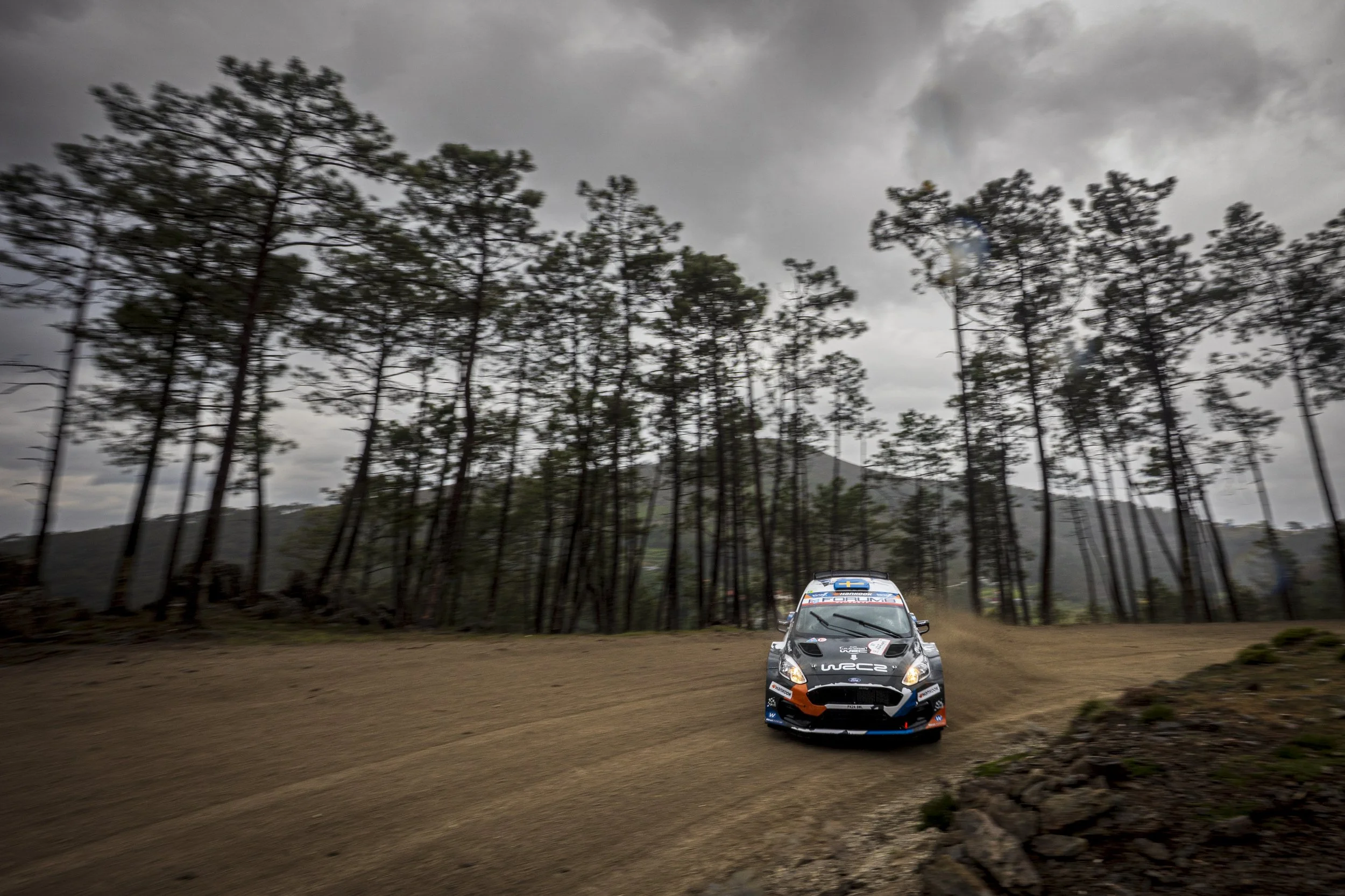Rally car racing on a dirt track in a forest, with trees and cloudy sky in the background.