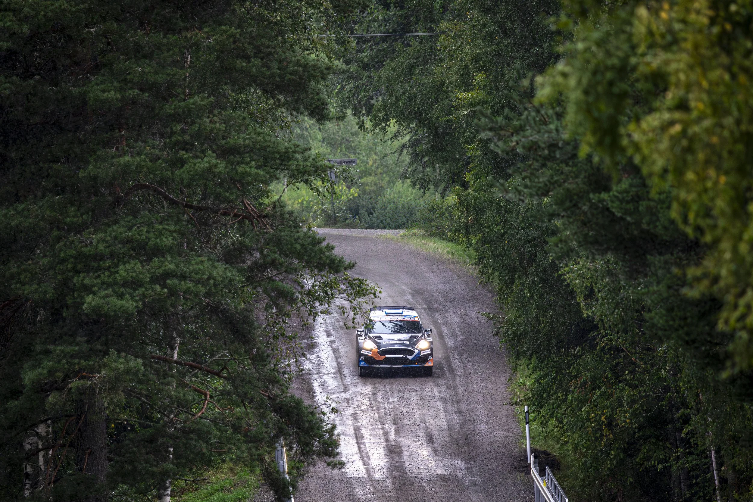 A rally car driving on a muddy, wet forest road surrounded by dense green trees during rainy weather.
