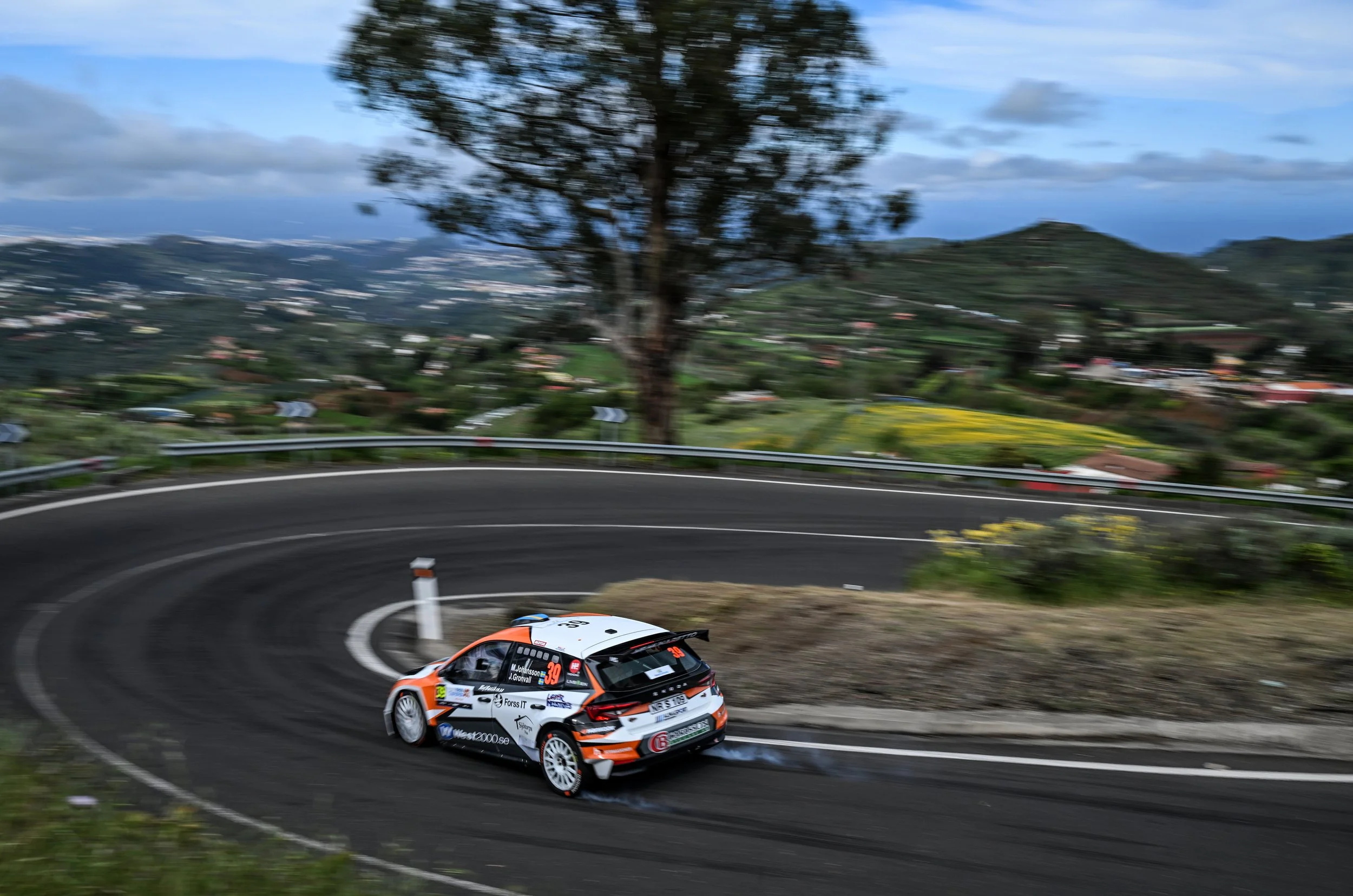 A rally car racing on a winding mountain road with a blurred background of trees and hills.