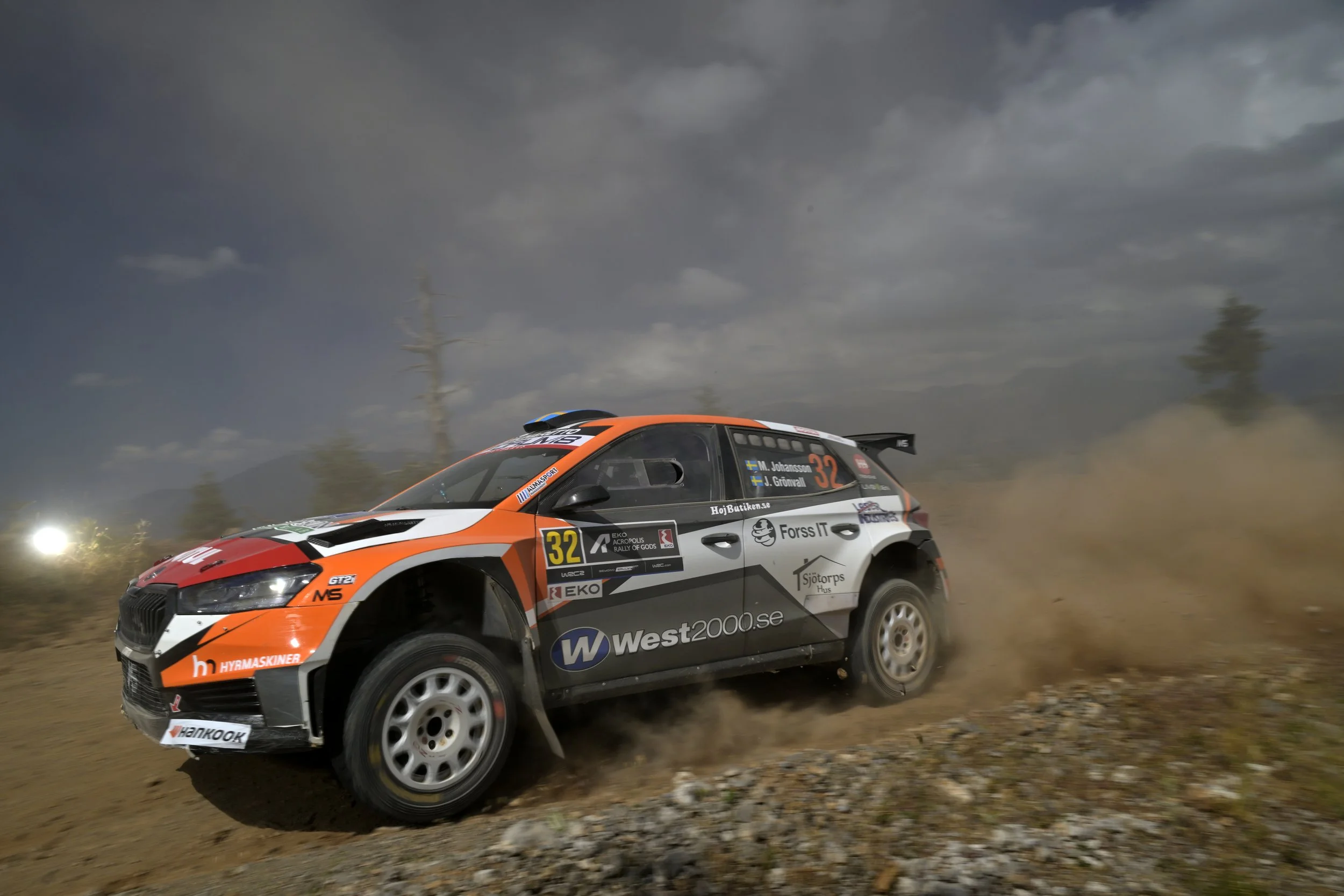 A rally car speeding on a dirt road with dust and debris kicked up behind it, under a cloudy sky.