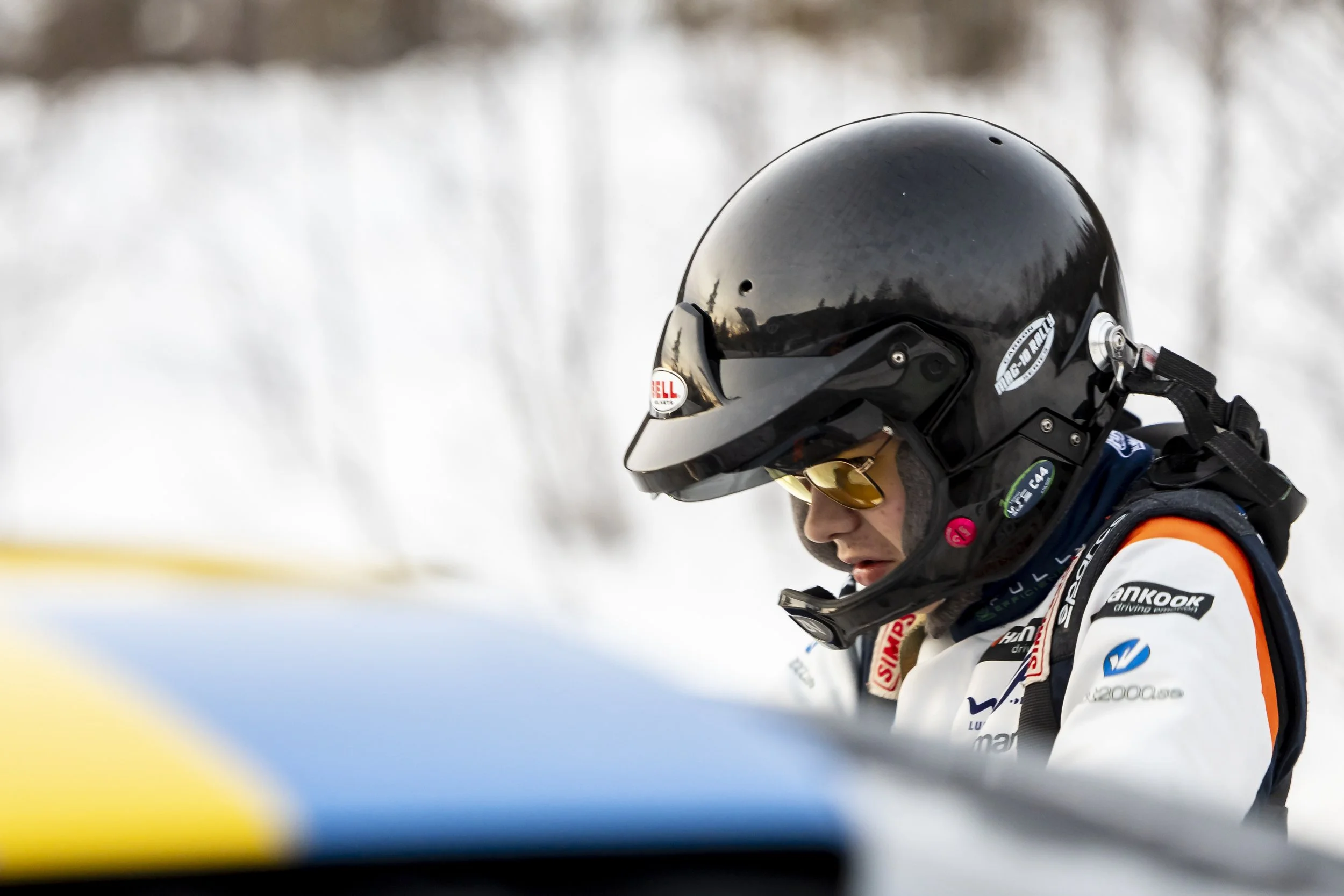 Close-up of a person in a racing helmet and gear, wearing reflective sunglasses, with snow and winter landscape in the background.