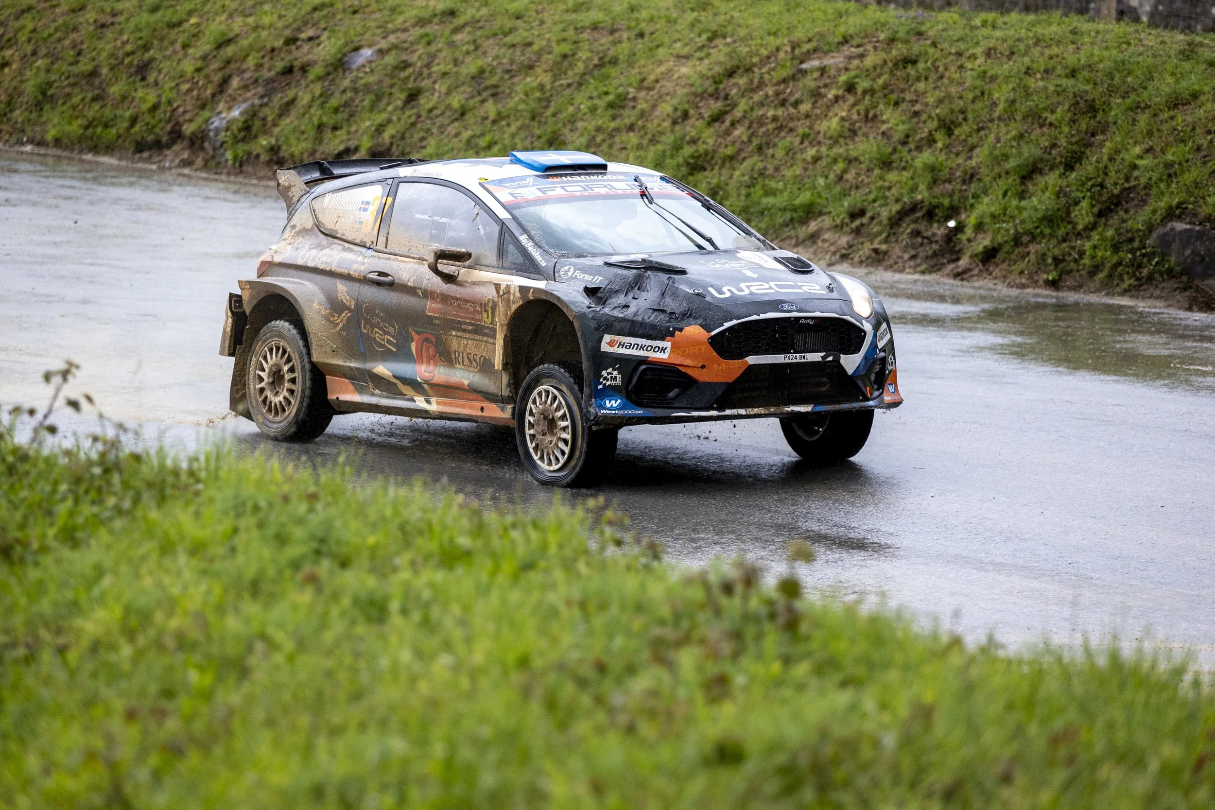 A rally car driving through a wet muddy terrain with water splashing around, surrounded by greenery.