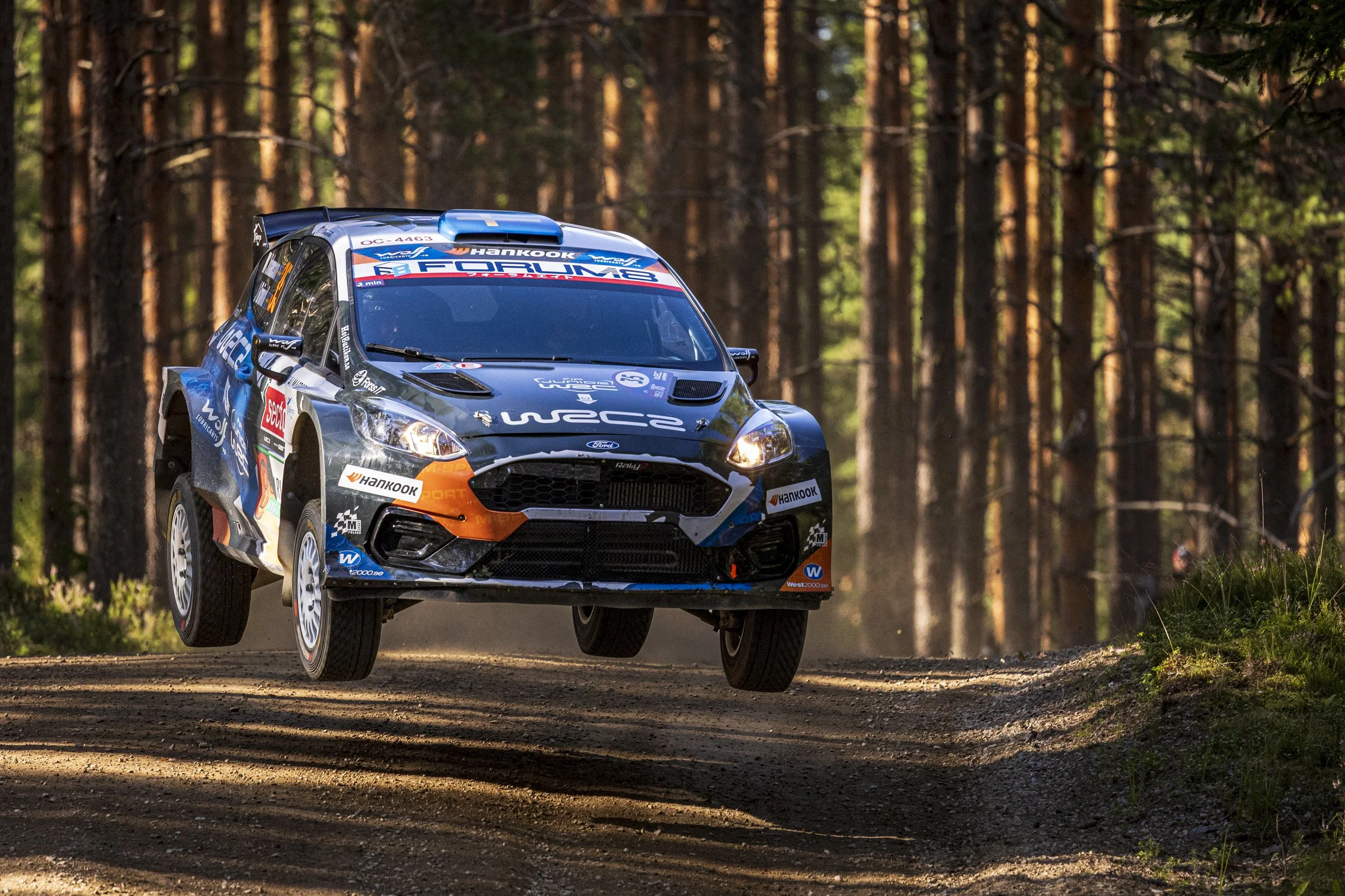 A rally car racing on a dirt forest trail with trees on both sides, kicking up dust as it speeds forward.