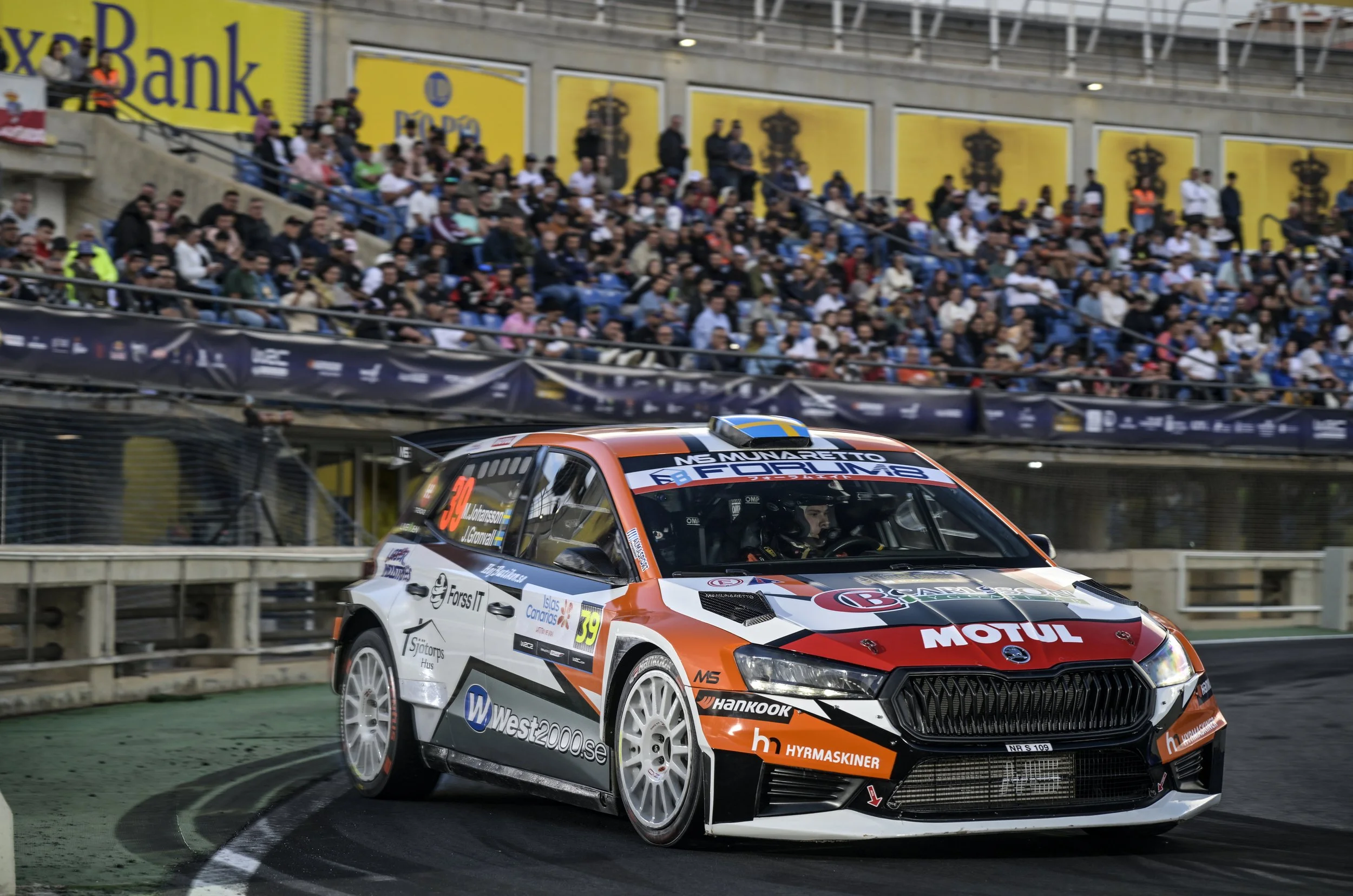 A rally car racing on a track with a crowd of spectators in the background, some staff and security personnel, and large advertisements on the stadium walls.