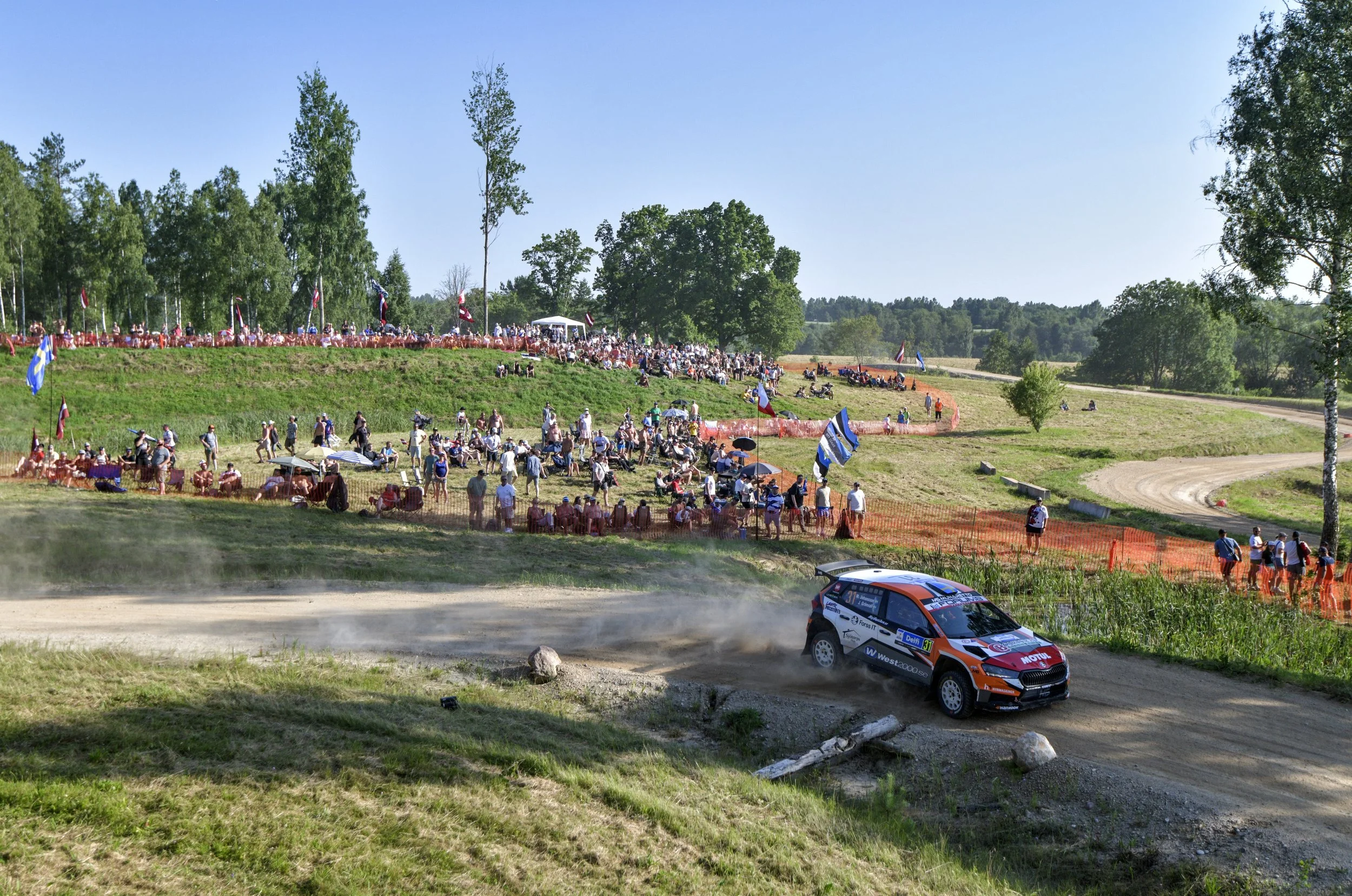 A rally car racing on a dirt track with dust rising behind it, surrounded by spectators and flags, in a rural area with trees and picnic areas.
