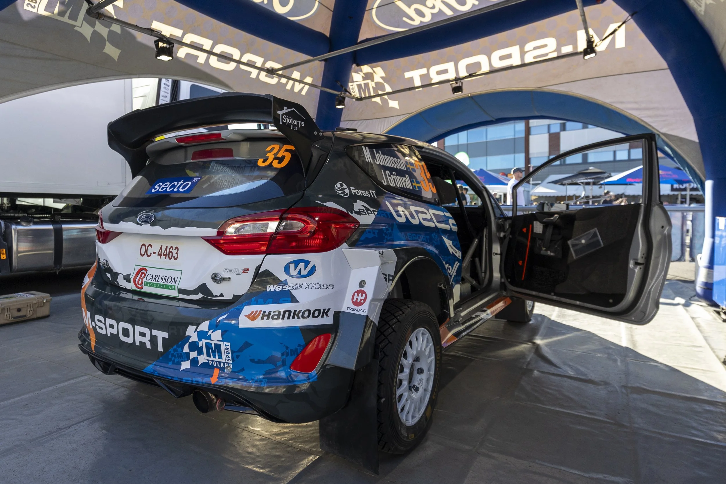 Race car with rally number 35, decorated with sponsor logos, partially inside a service tent at a racing event. The tent has a blue and beige canopy with writing visible on the inside.