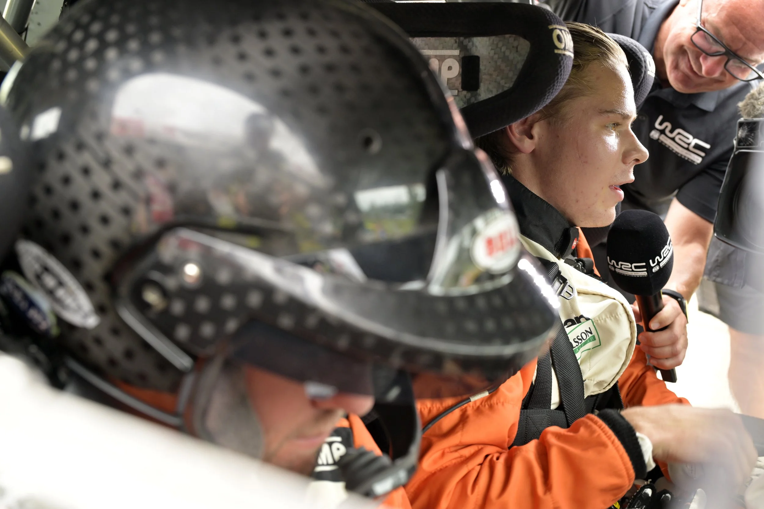 A close-up of a racing helmet in the foreground, with a race car driver and a team member in racing gear having a conversation in the background.