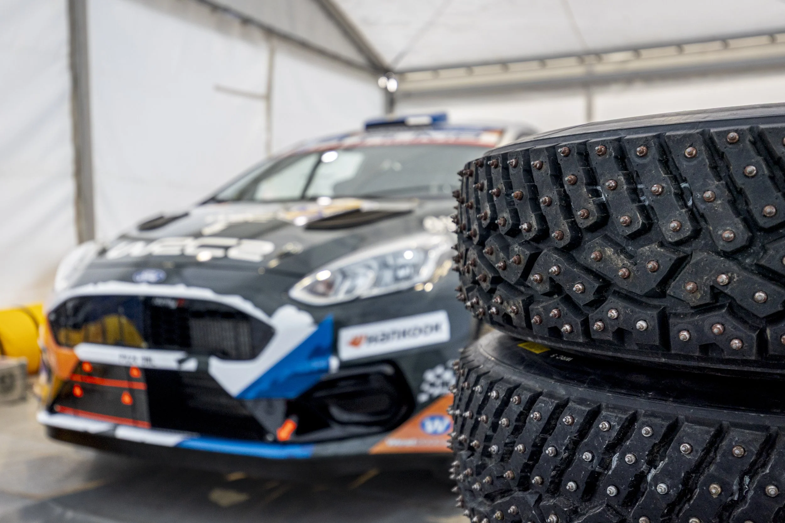 Close-up of a stack of black racing tires with metal studs, with a blurred race car in the background inside a garage or service area.