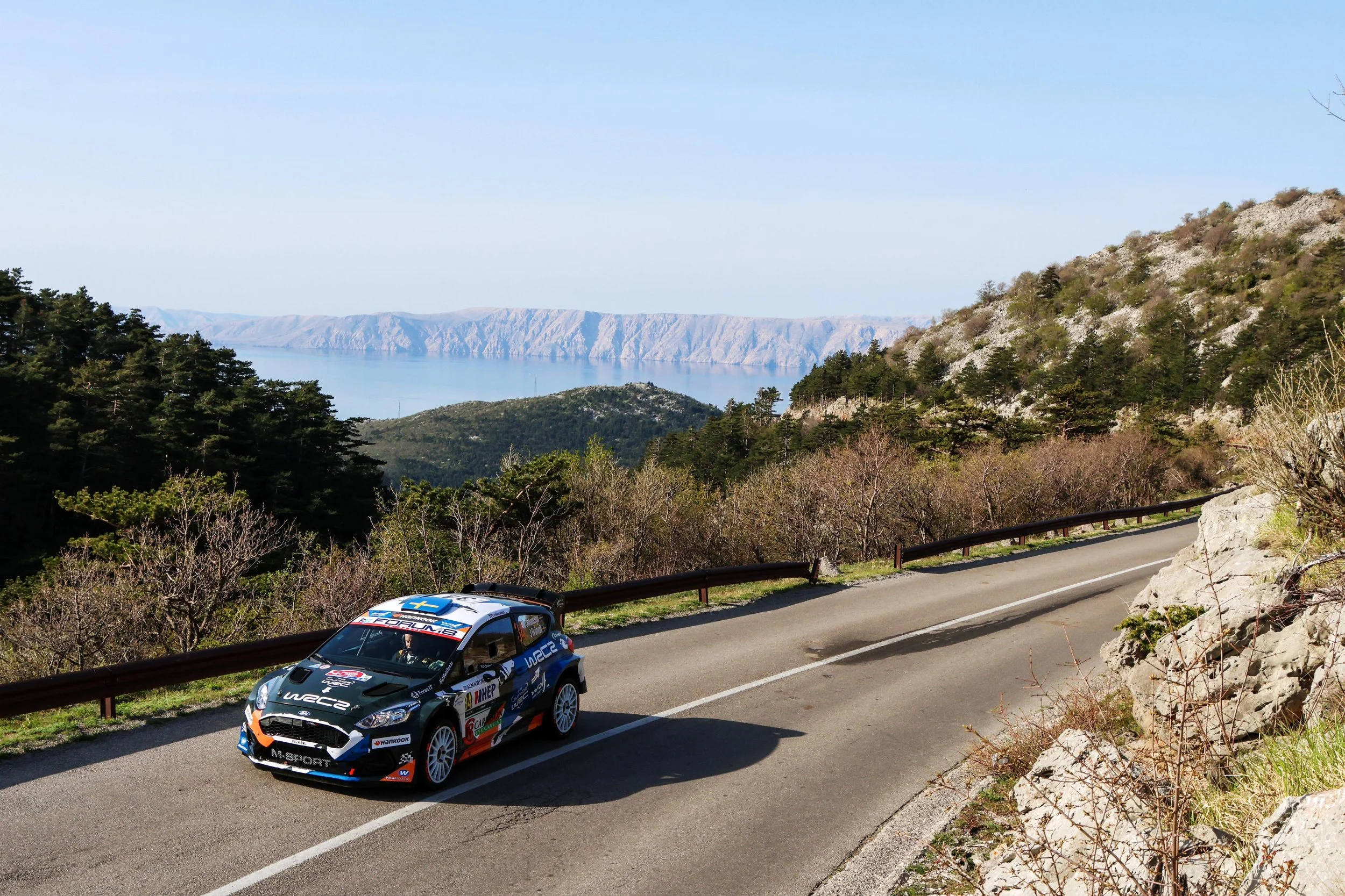 A rally car driving on a winding mountain road with a guardrail, surrounded by trees and rocks, overlooking a body of water and distant mountains.