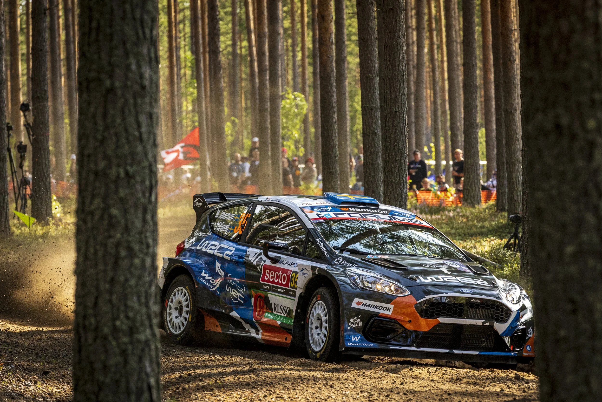 Race car driving through a forest during a rally race, with spectators and event flags in the background.