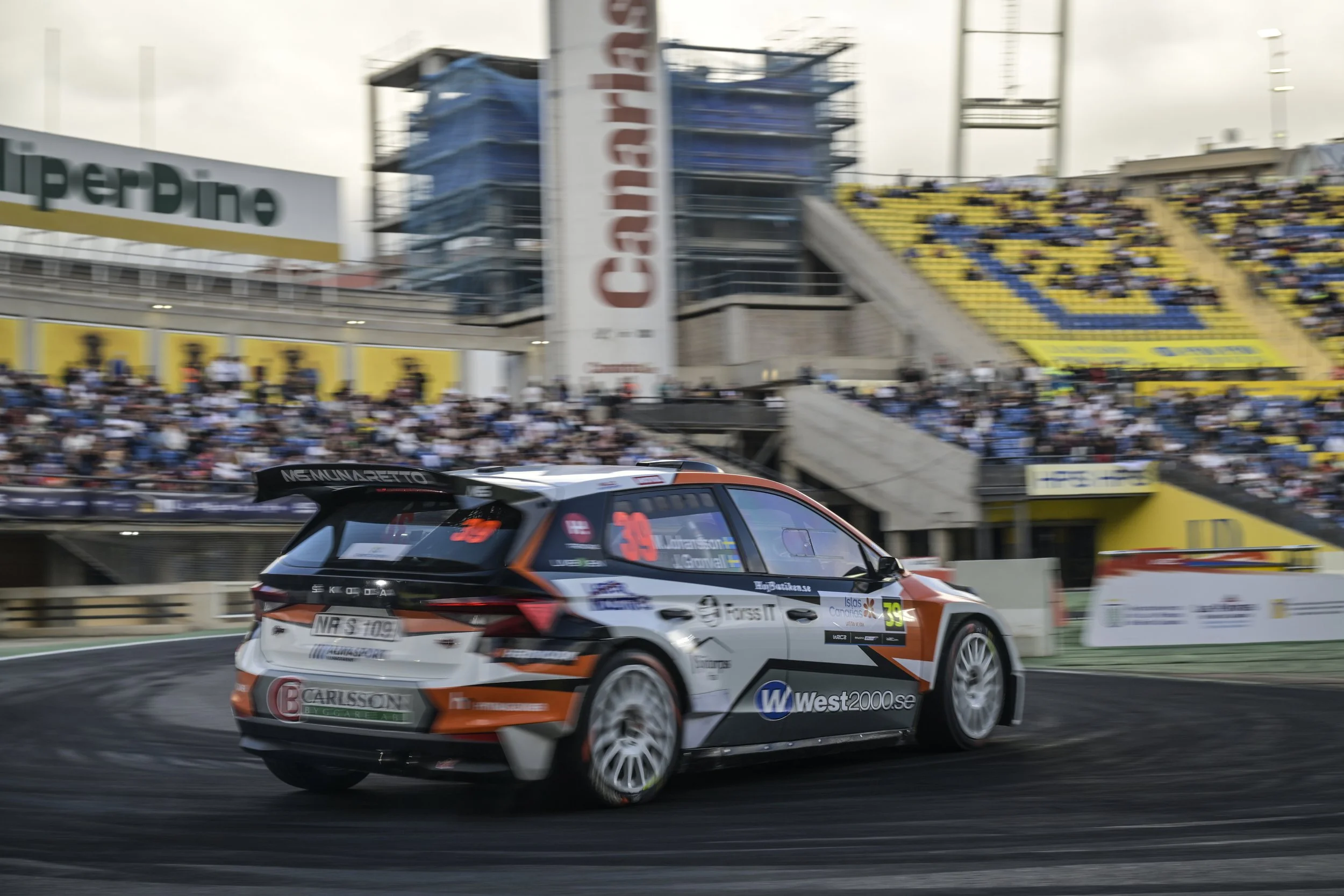 A rally car racing on a track during a motorsport event with spectators seated in the grandstands in the background.