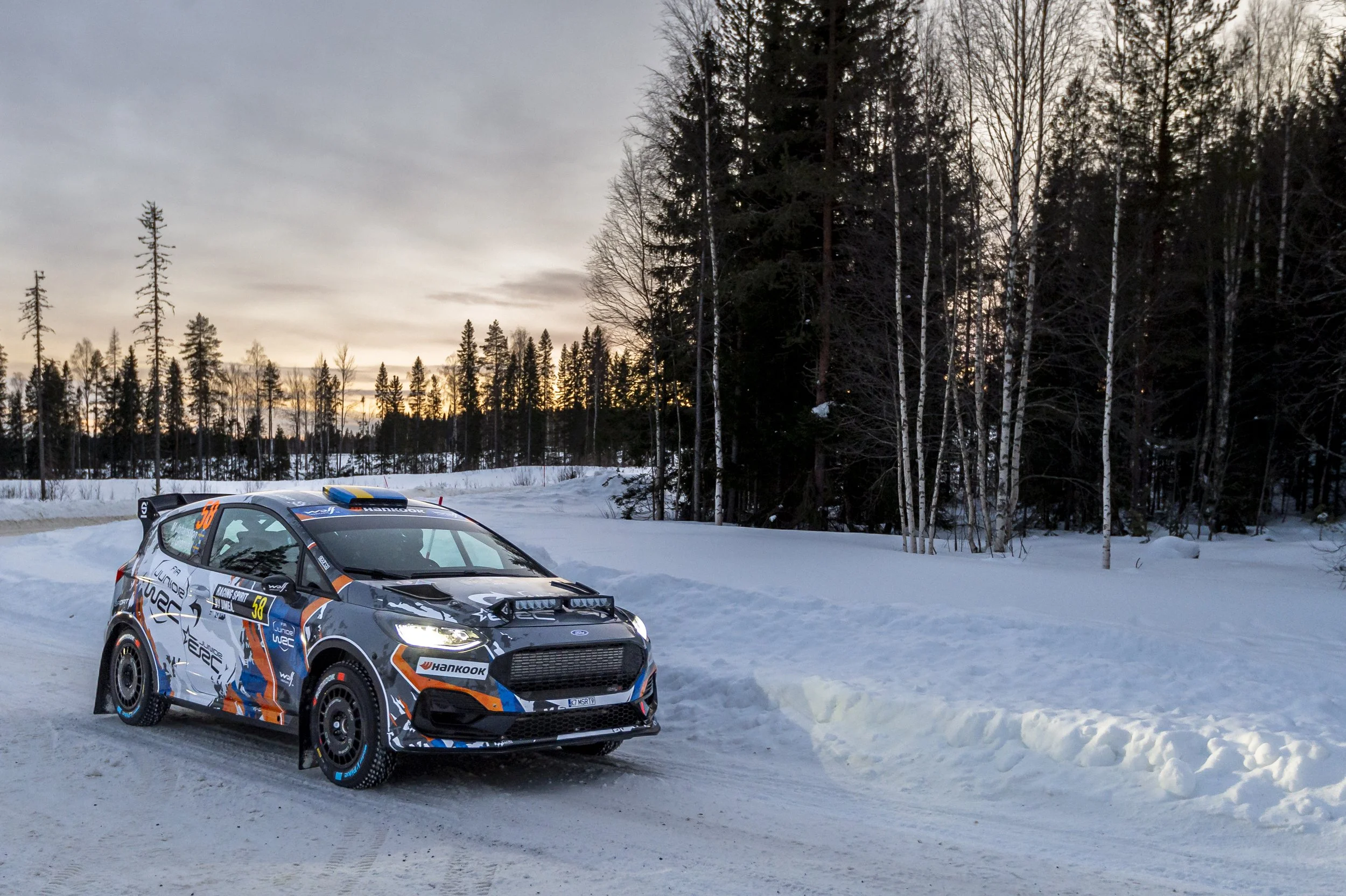 A rally car on a snow-covered road with a forest of tall, thin trees in the background and a cloudy sky at sunset.