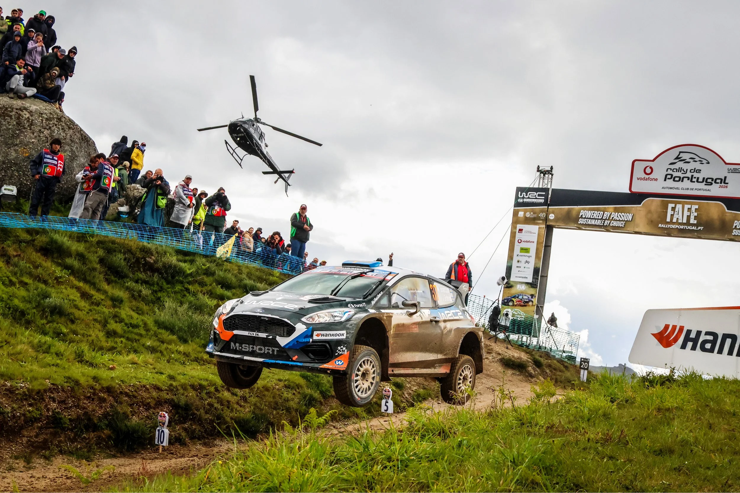 Race car driving on a dirt track with spectators watching from a hillside and a helicopter flying overhead at the rally de Portugal.