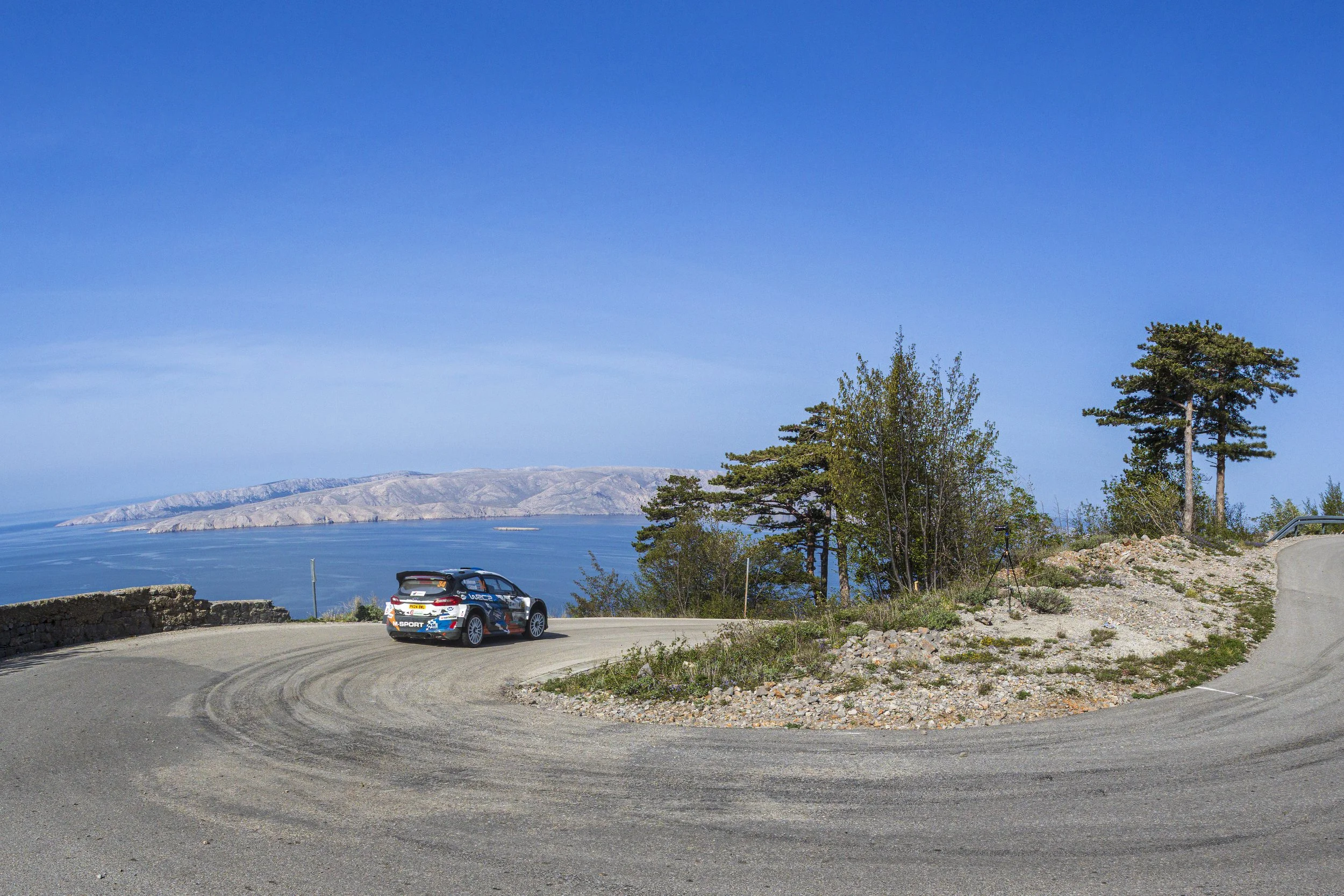 A rally car driving on a curved mountain road with a scenic view of water and mountains in the background, surrounded by trees and rocky terrain.
