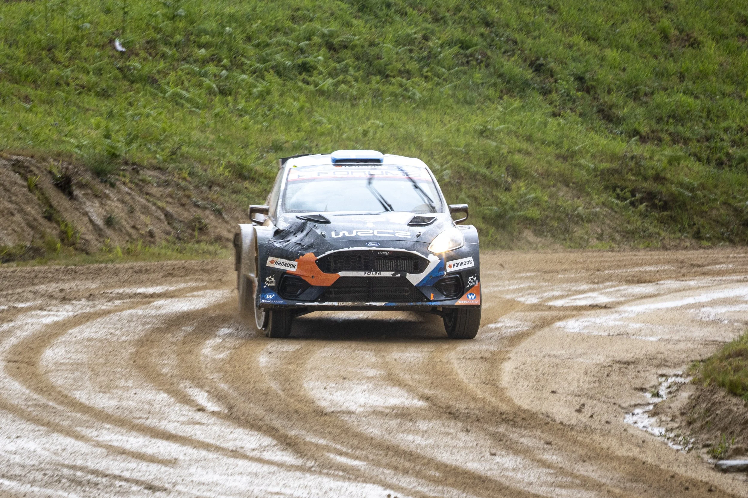 A rally car speeding on a dirt track with tire tracks, surrounded by grassy landscape.
