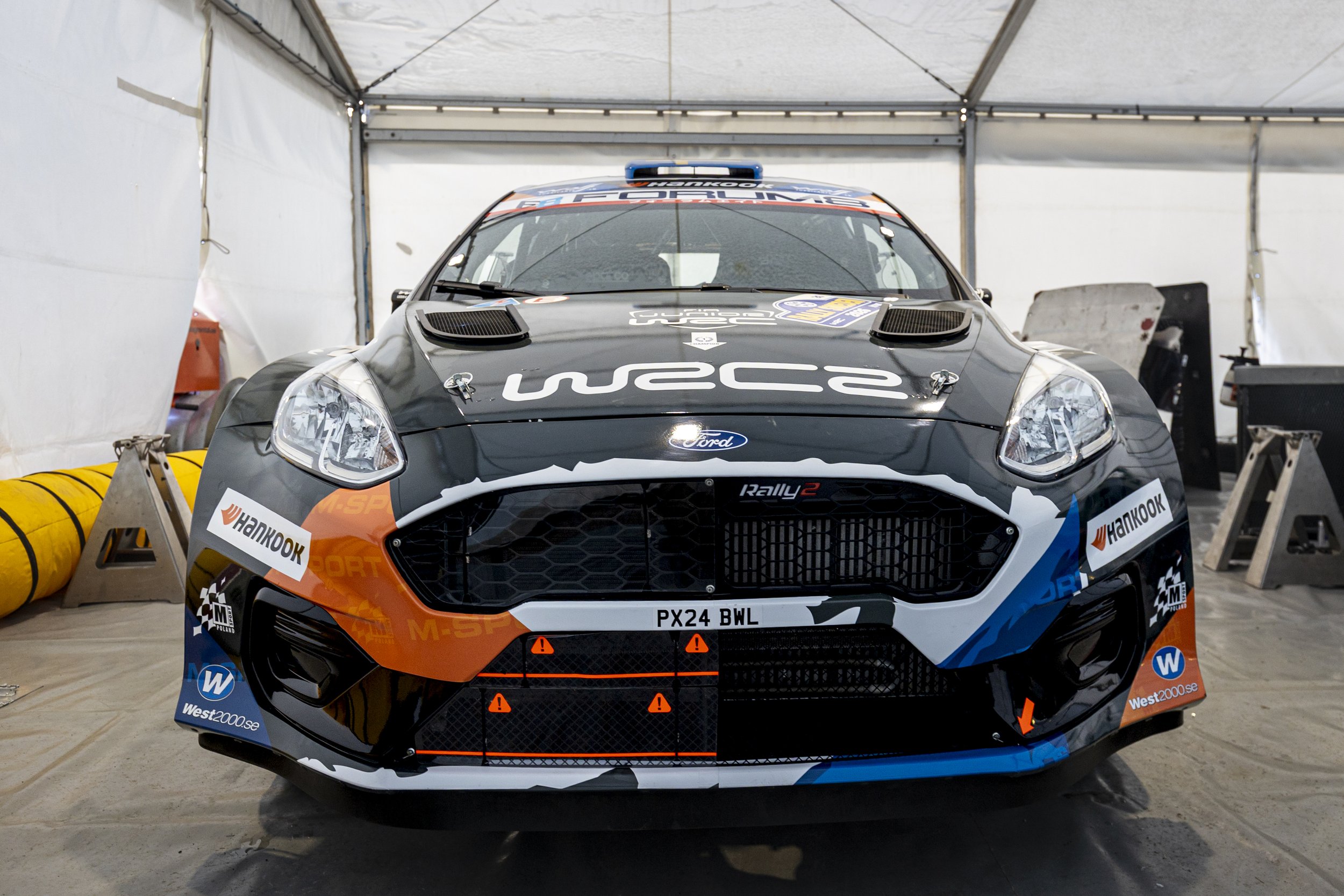 Front view of a black and orange rally car inside a tent, with various sponsor logos and a license plate reading PX24 BWL.